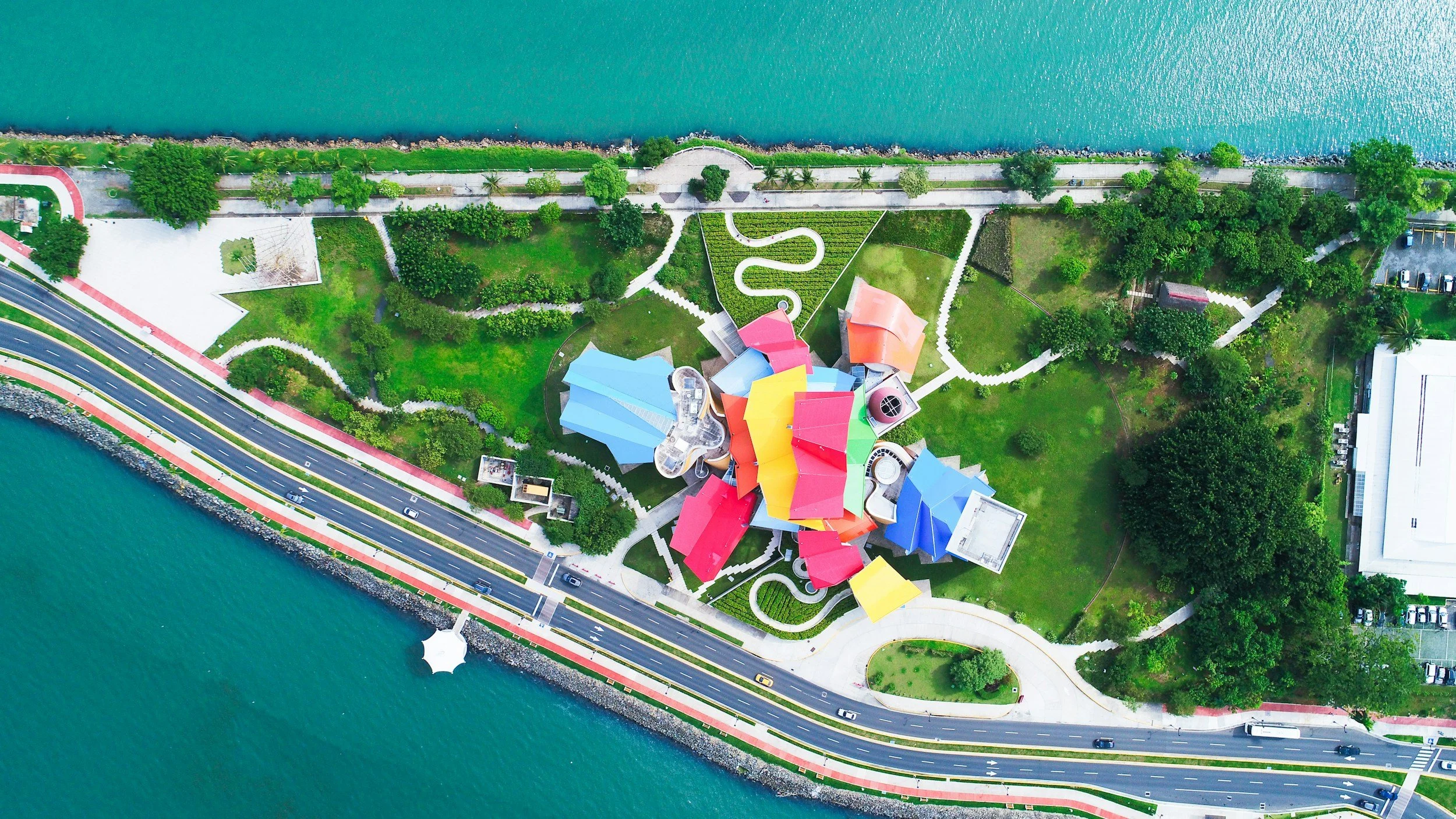 Aerial view of a colorful oceanfront park with multicolored buildings, walking paths, green lawns, trees, and nearby roads and water.
