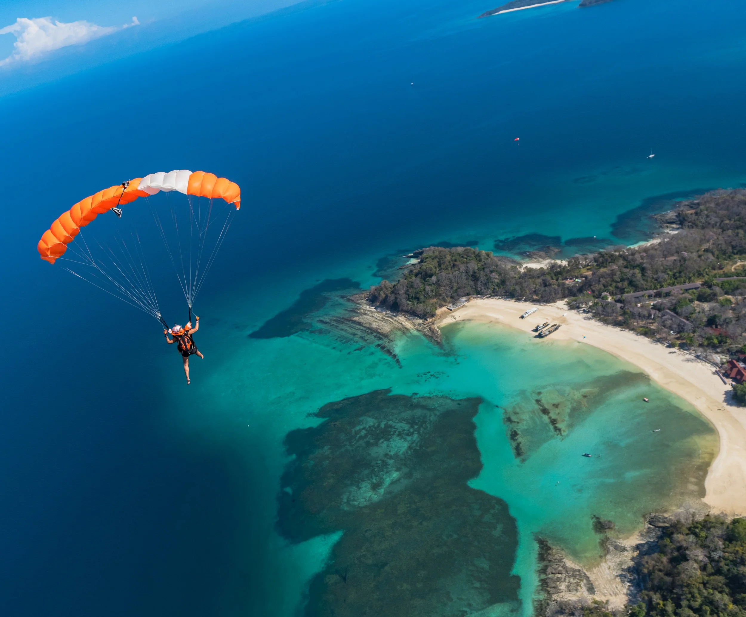 A person paragliding over a turquoise coastal area with a sandy beach, lush green trees, and a bright blue ocean under a partly cloudy sky.