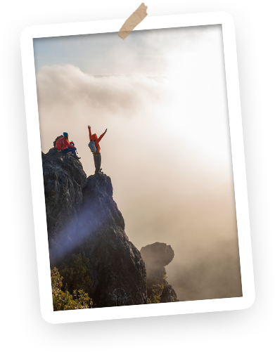 Two hikers standing on a rocky mountain peak, celebrating with arms raised, surrounded by fog and clouds.