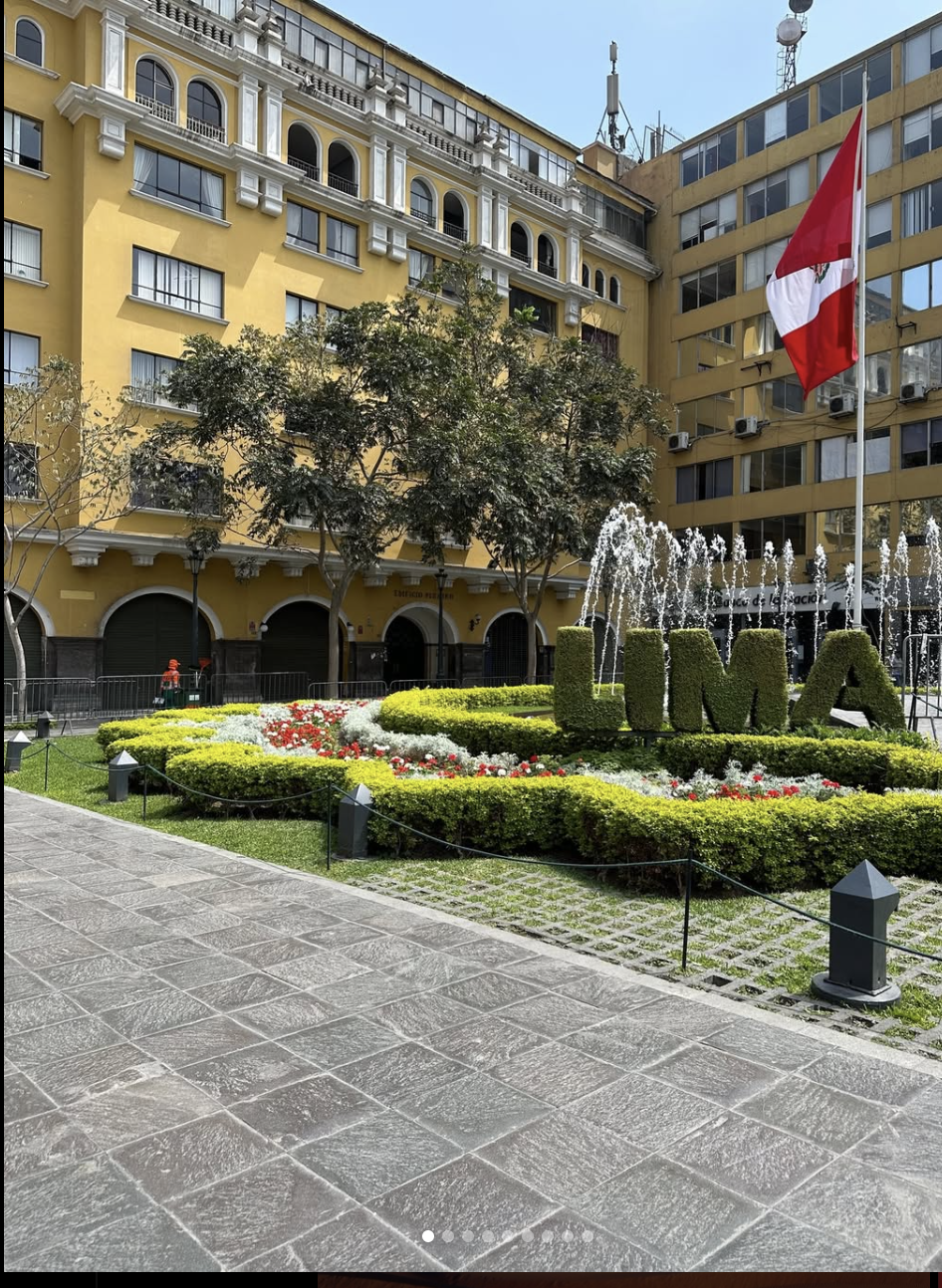 A city square with a yellow building in the background, decorated with arched windows and balconies, and a flagpole with a Peruvian flag. There is a flower bed with the word 'LIMA' made of greenery, surrounded by small fountains and decorative lights. The scene is sunny with a clear blue sky.