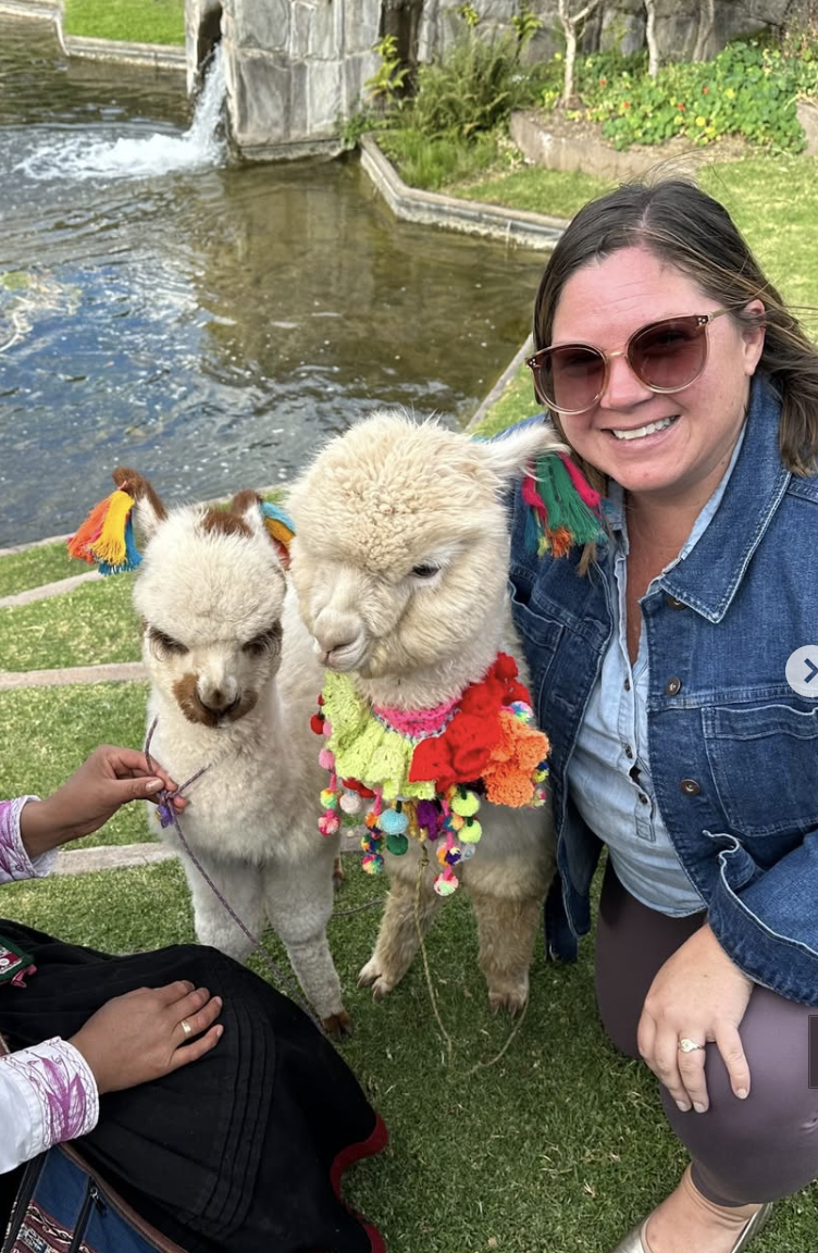 A woman smiling beside two decorated alpacas near a water feature in a park.