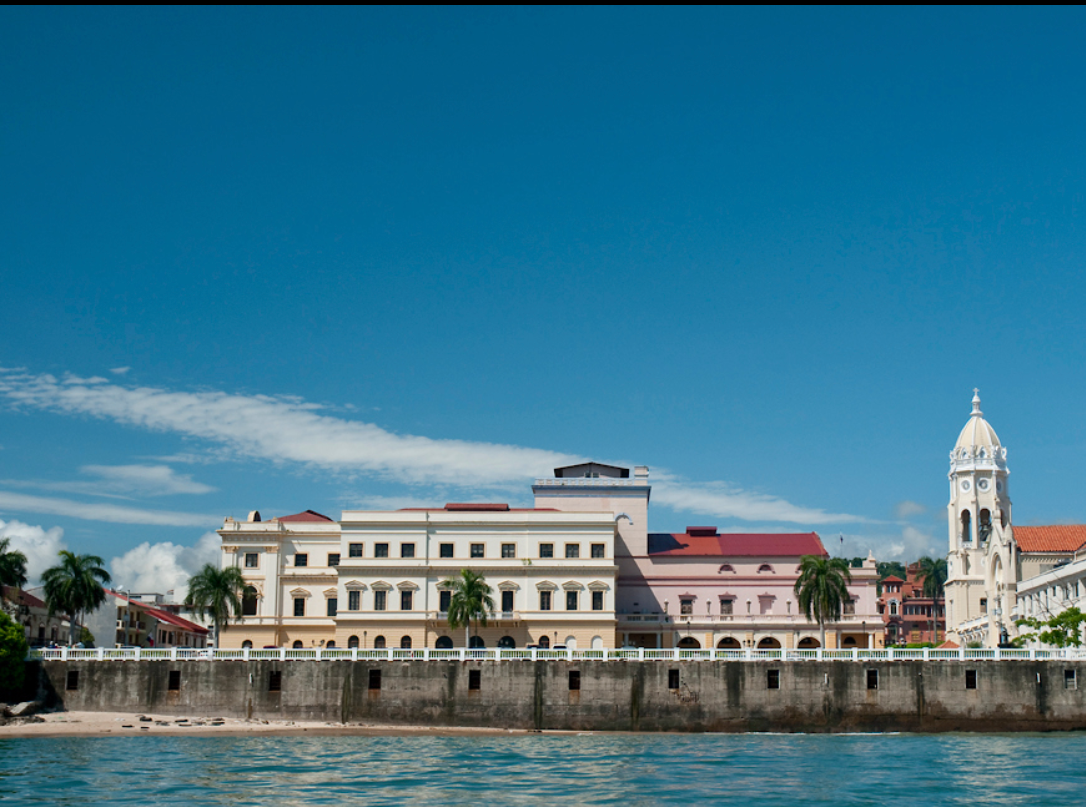 Buildings on a waterfront with a concrete seawall, palm trees, and a blue sky with clouds.