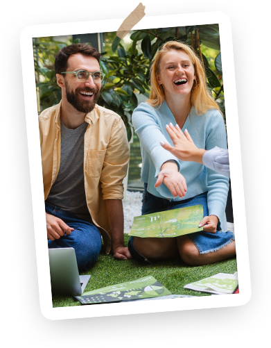 Two people sitting on the grass indoors, laughing and playing a game with a person off-camera, surrounded by plants and greenery.
