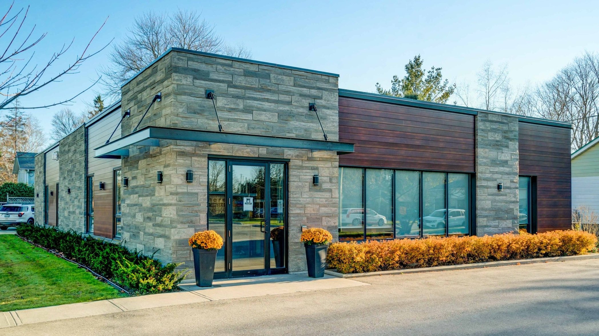Modern commercial building with stone and wood exterior, large glass windows, and potted chrysanthemums near the entrance, surrounded by landscaping and parked cars.