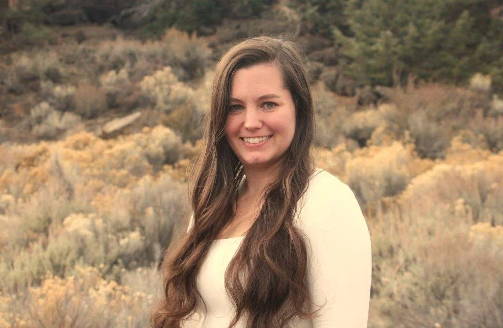 A person with long dark hair wearing a black top and dangling earrings, smiling in front of a leafy green background.