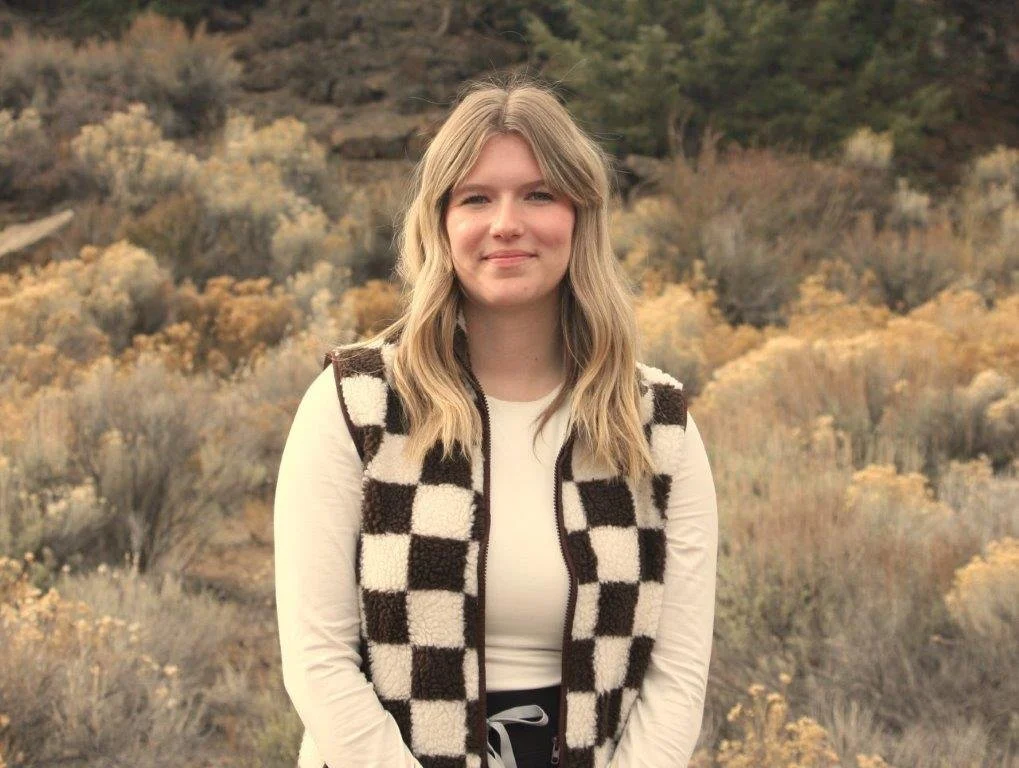 Young woman with blonde hair smiling, wearing a black top and a necklace with a star pendant, with a blurred green background.