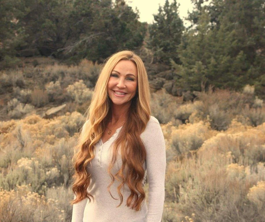 Smiling woman with long hair wearing a floral dress and necklace, set against a background of green leaves.