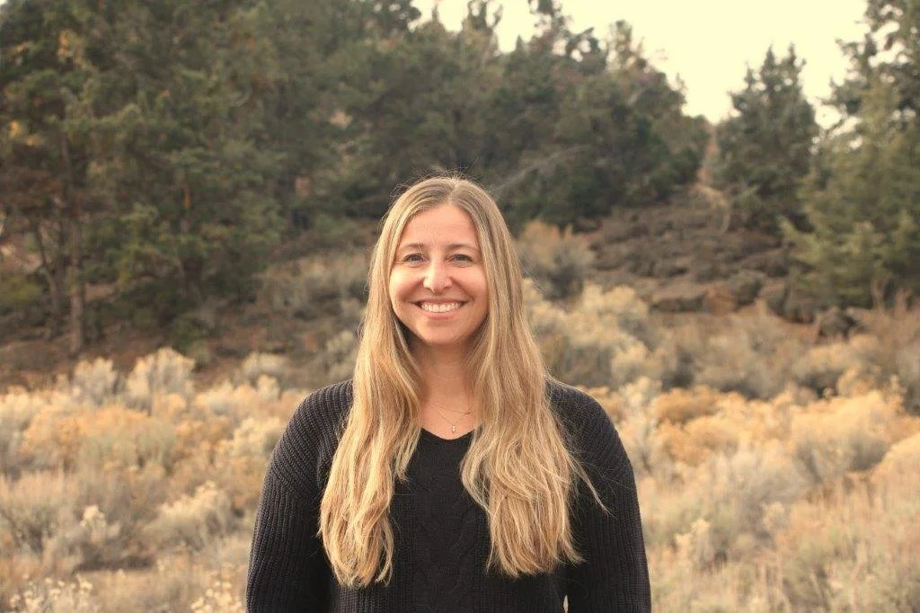 Woman with long blonde hair smiling, wearing a black top, green foliage in the background.