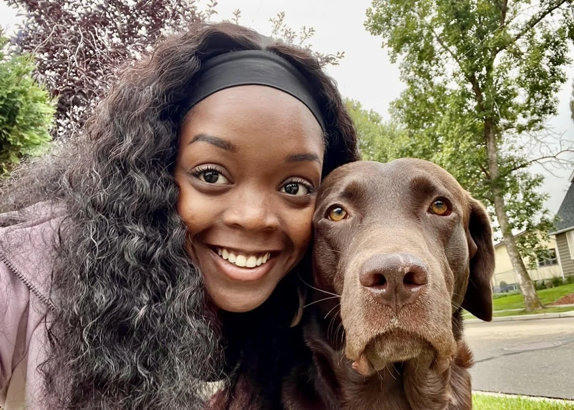 Smiling person with curly hair taking a selfie with a brown Labrador dog outdoors.