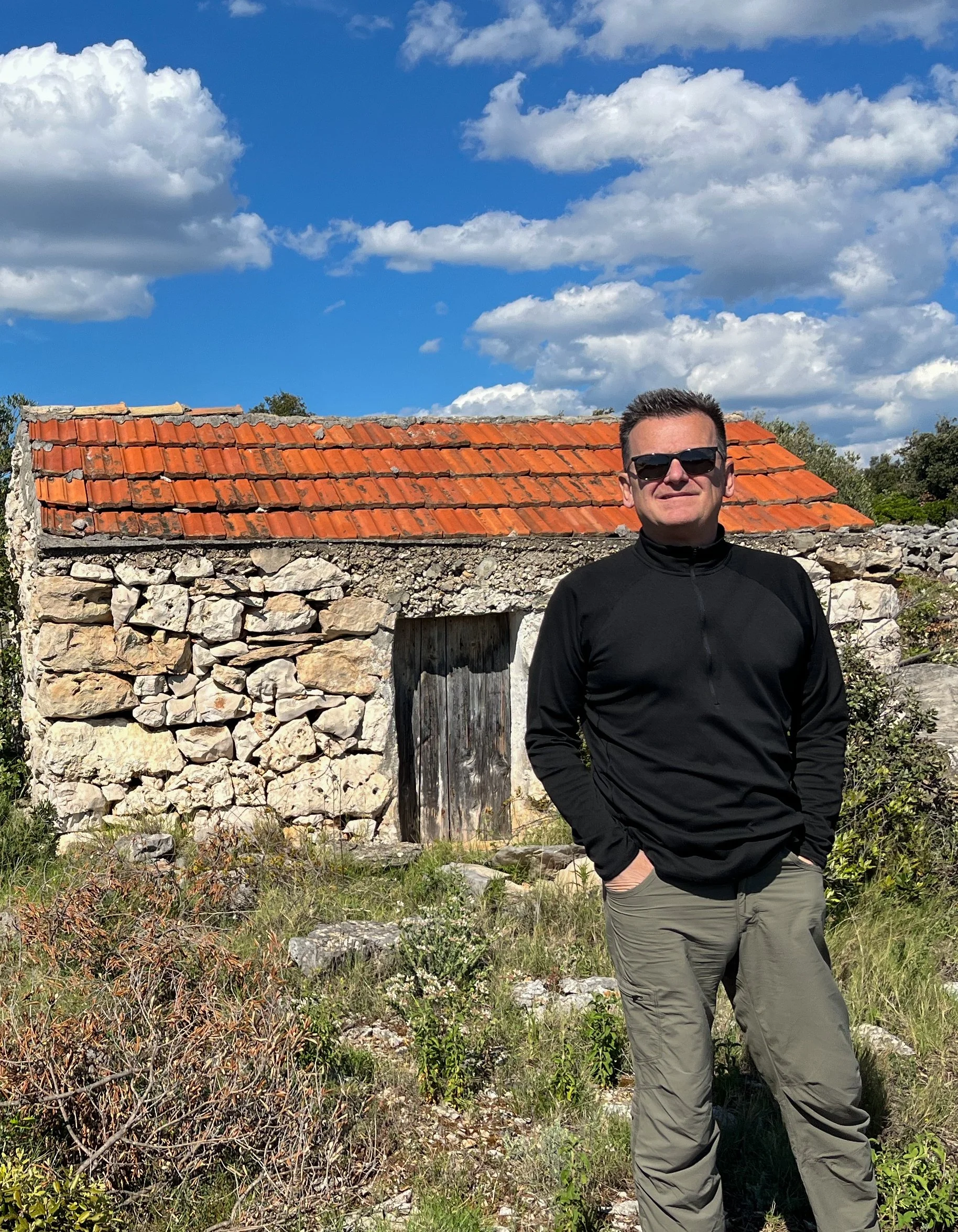 A man wearing sunglasses, a black jacket, and gray pants standing outdoors in front of an old stone building with a red-tiled roof under a partly cloudy sky.