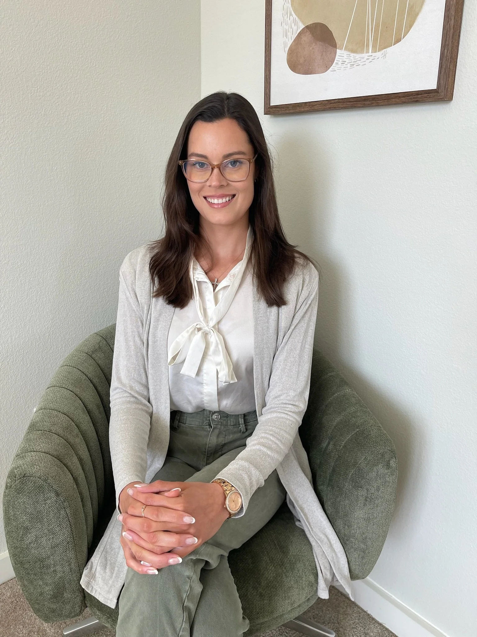 A woman with glasses and long brown hair sitting on a green armchair, smiling, in a room with beige walls and a framed abstract art piece hanging above her.