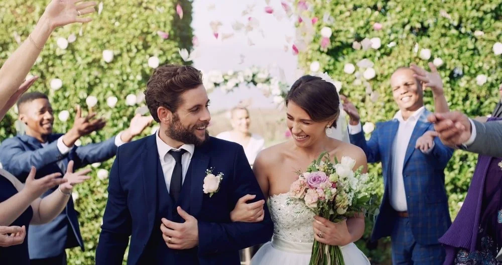 A bride and groom walking together at their outdoor wedding surrounded by friends and family celebrating with confetti and smiling.