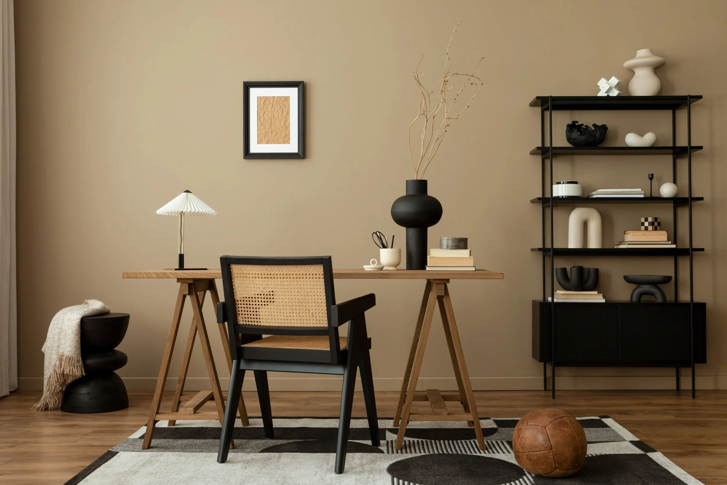 Modern living room with a wooden desk, black and beige chair, black accents, decorative vases, books, and a black shelving unit against a beige wall.
