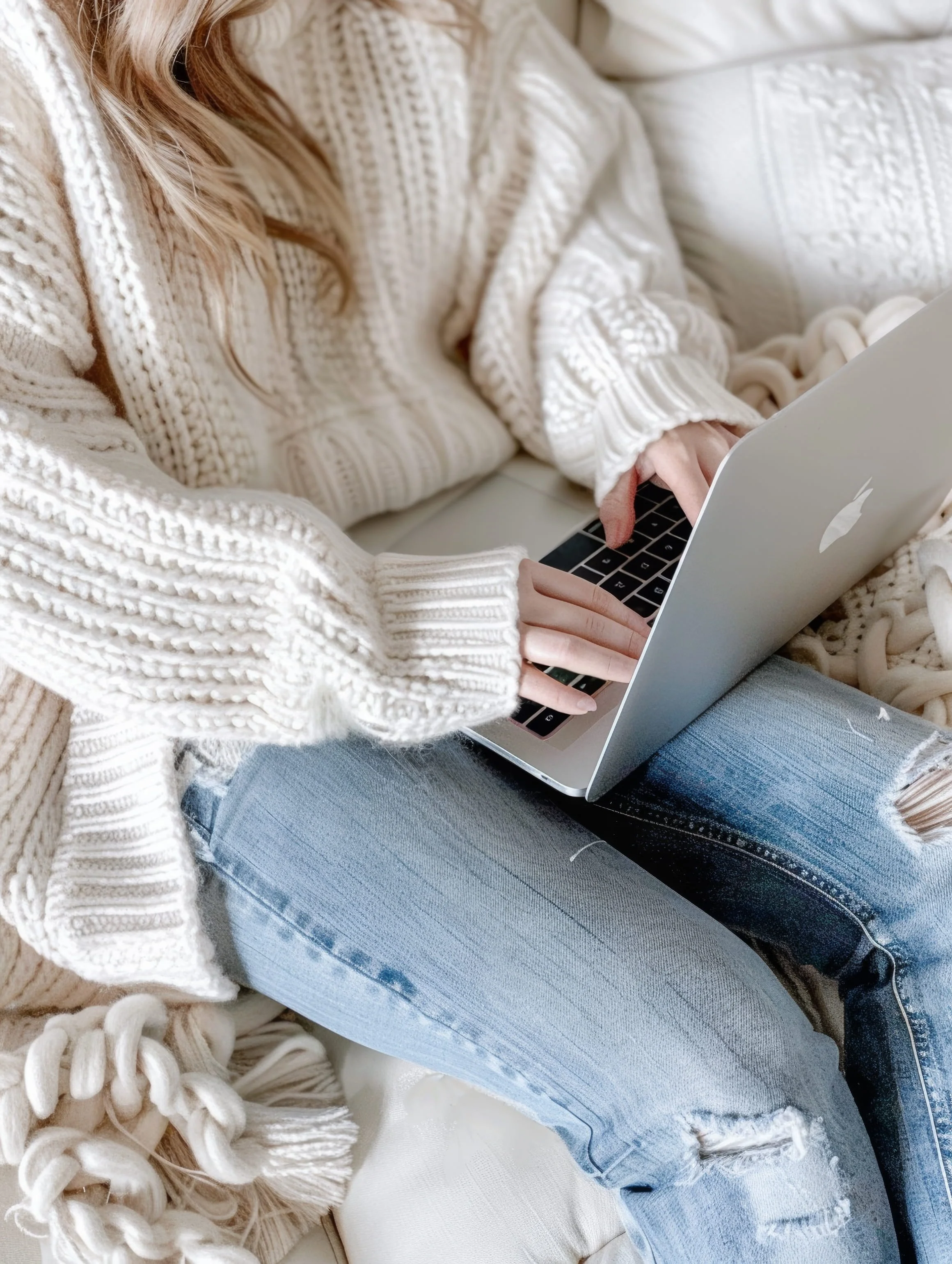 Person sitting on a white couch wearing a chunky knit beige sweater with long sleeves, light blue ripped jeans, and using a silver MacBook.