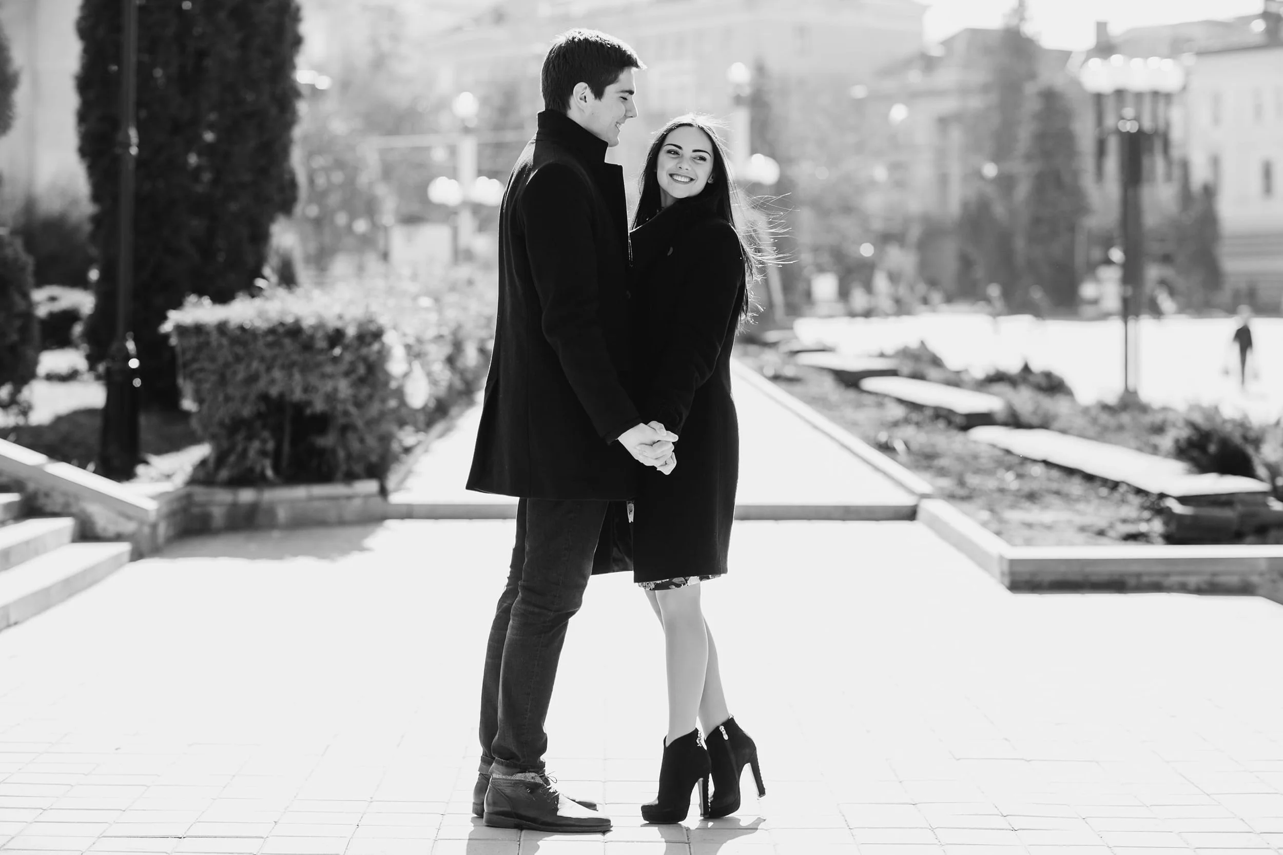 A smiling young couple holding hands and looking at each other outdoors on a sunny day, with trees and buildings in the background.
