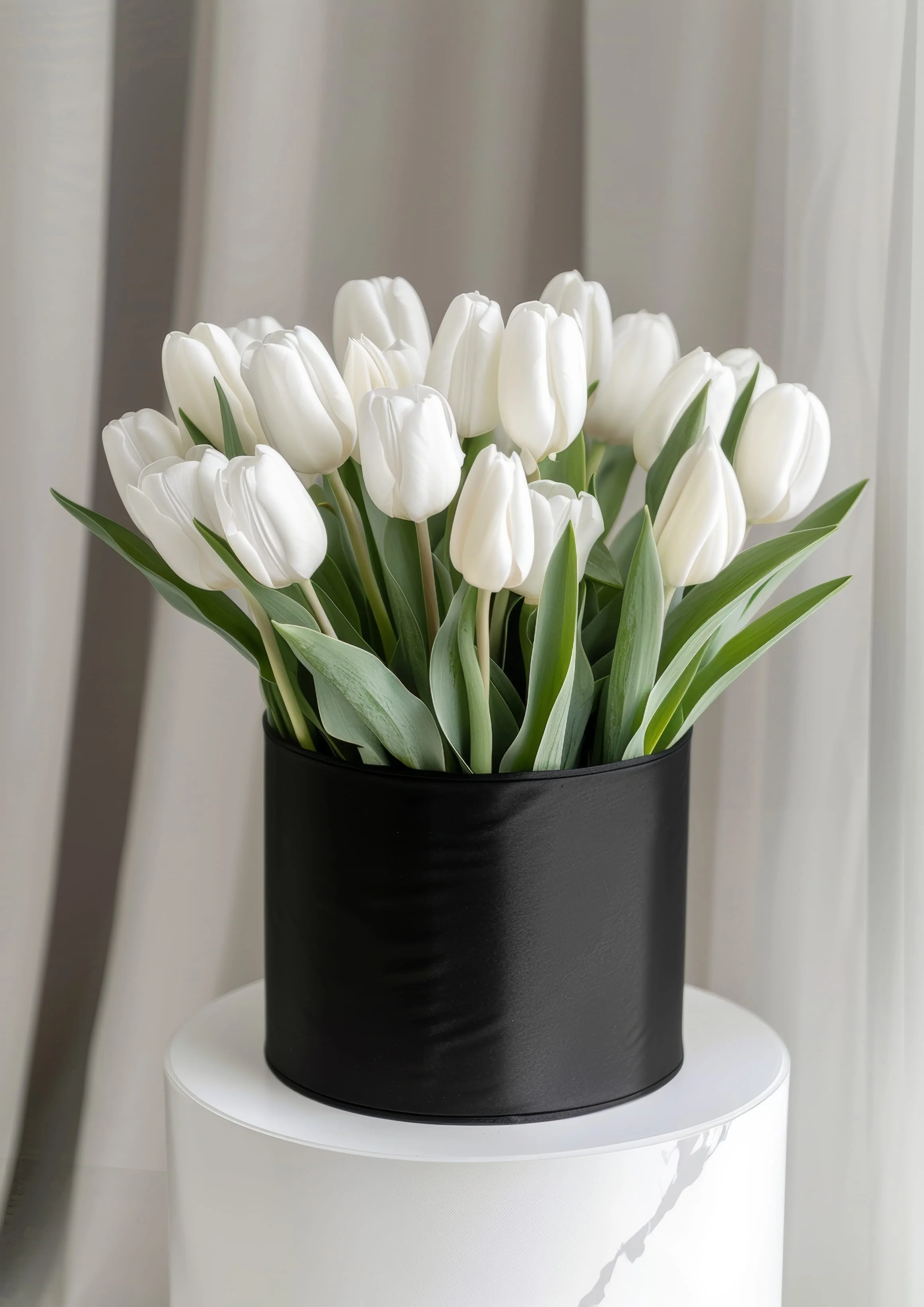 White tulips in a black vase on a round white table, with a white curtain background.