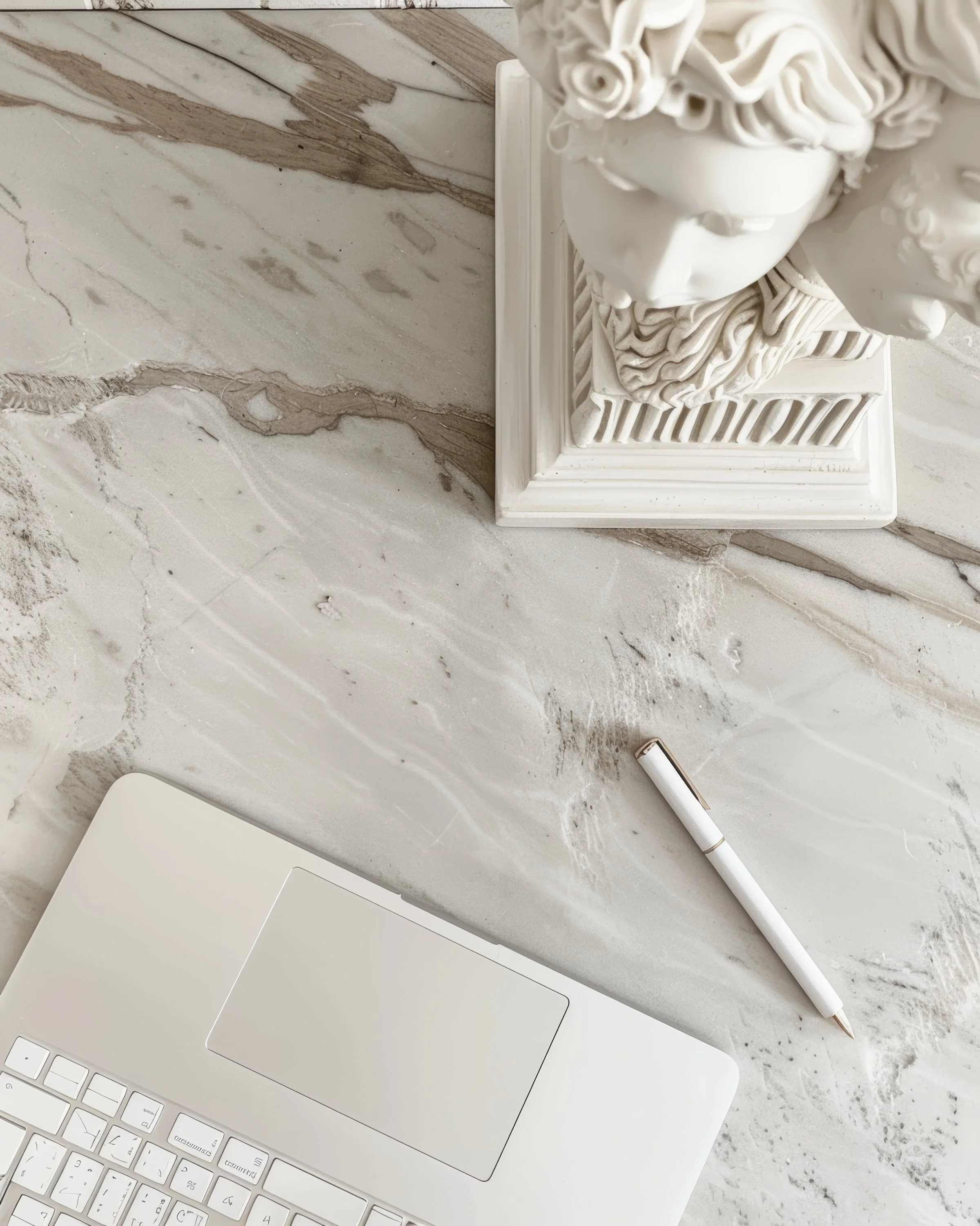 A marble countertop with a white decorative bust sculpture featuring curly hair and a detailed base, next to a silver laptop, a white pen, and part of a white keyboard.