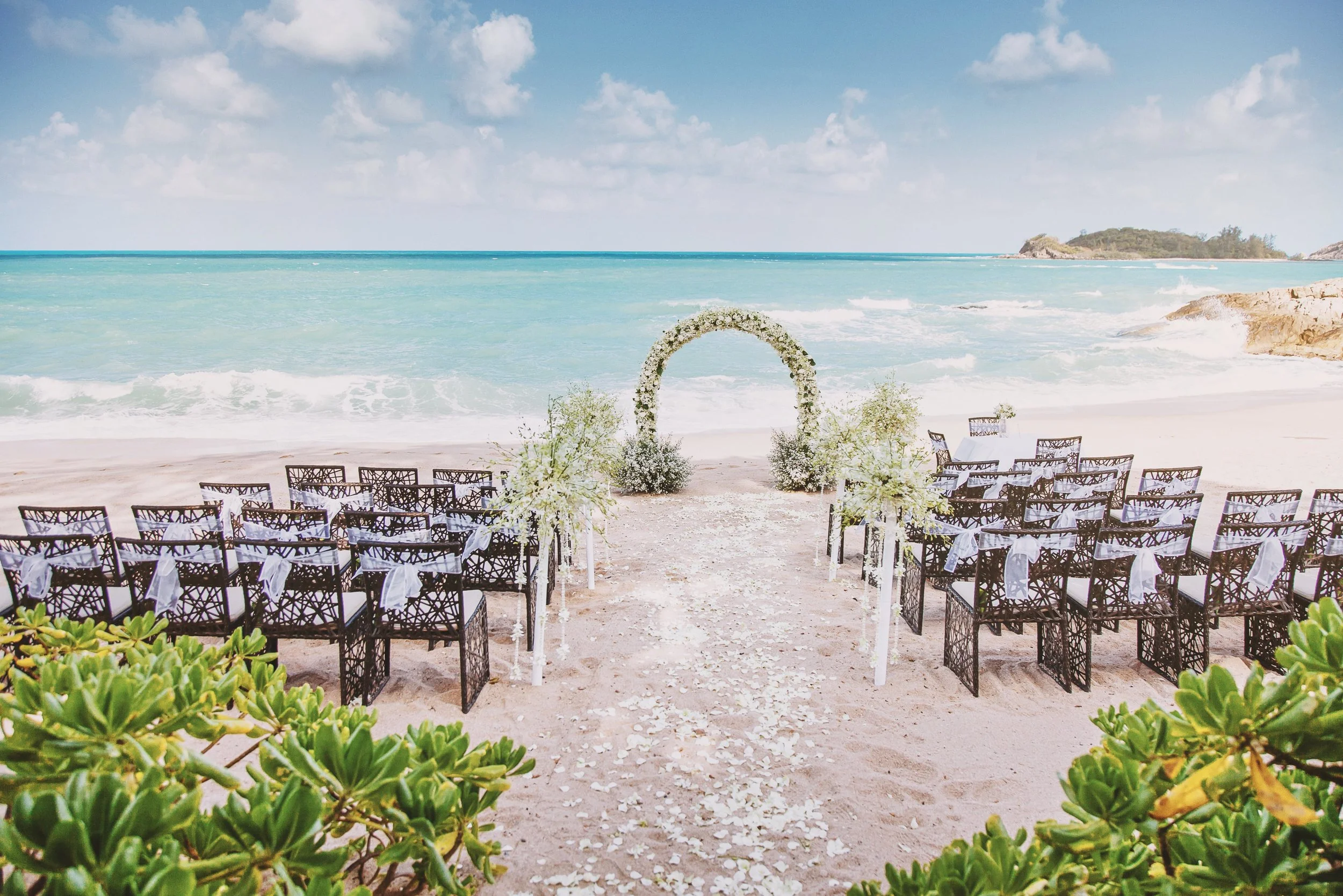 Beach wedding setup with chairs, a floral arch, and ocean view.