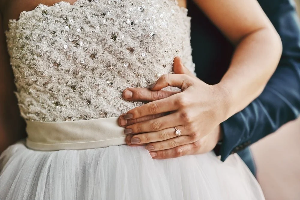 Close-up of a bride's hand with an engagement ring resting on her waist, showing her wedding dress with beaded embellishments and a satin waistband, and a groom's arm wrapped around her.