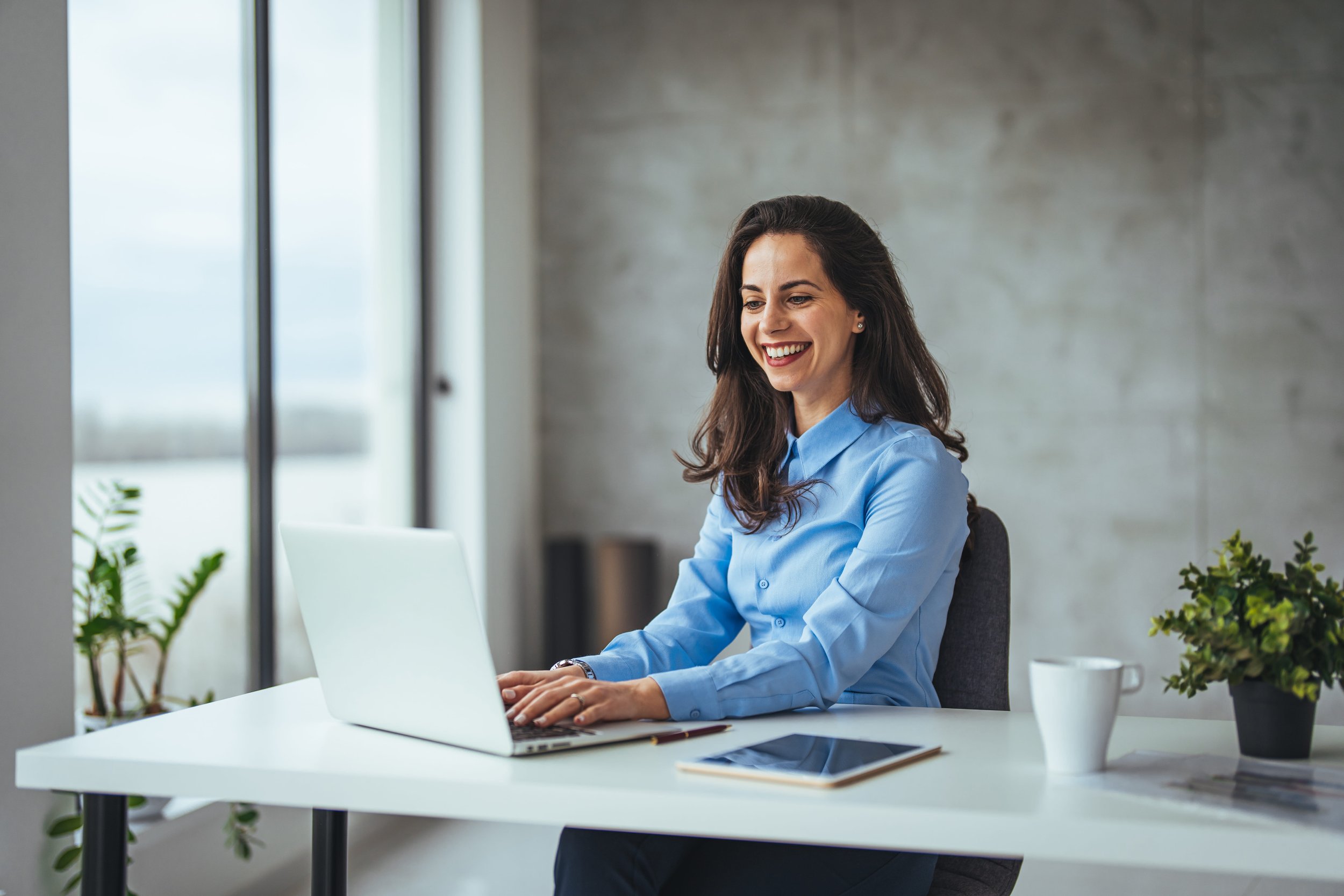 Woman with long dark hair in a light blue shirt sitting at a white desk, smiling while working on a silver laptop in a modern office with large windows and plants.