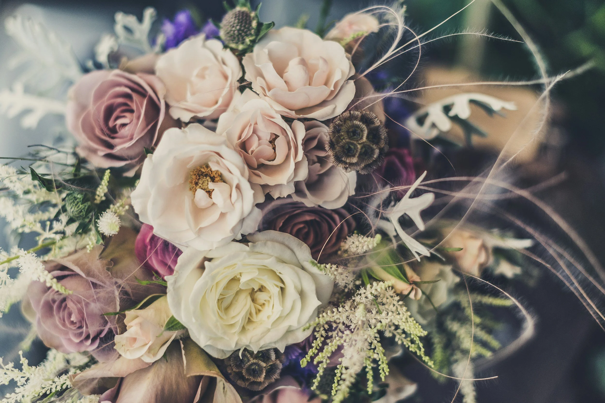 A close-up of a bouquet of flowers featuring roses, lisianthus, and other blooms in shades of white, blush, and purple, accented with greenery and decorative wispy elements.