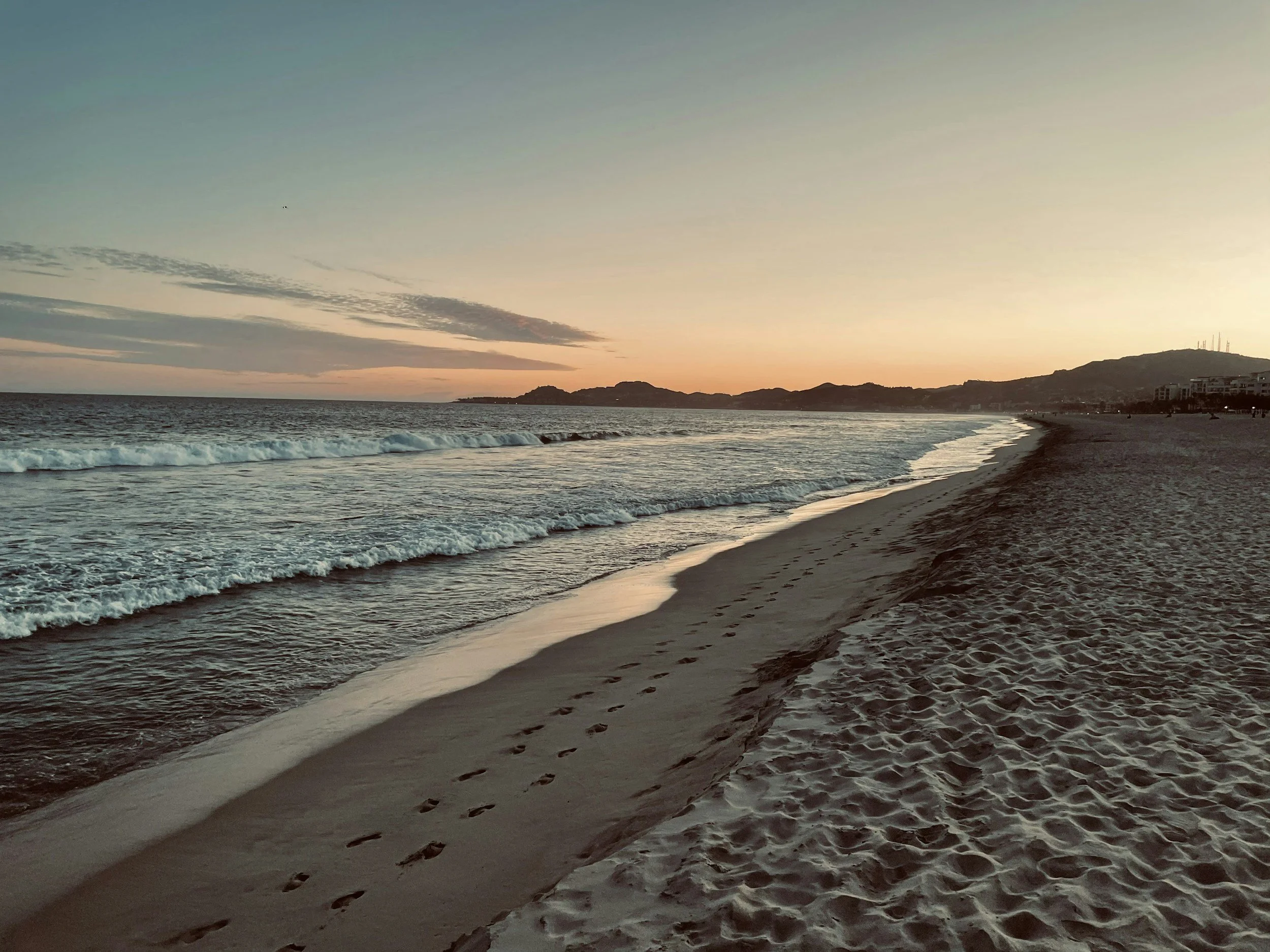 A tranquil beach at sunset with footprints in the sand, gentle waves washing ashore, distant hills, and a mostly clear sky with some clouds.