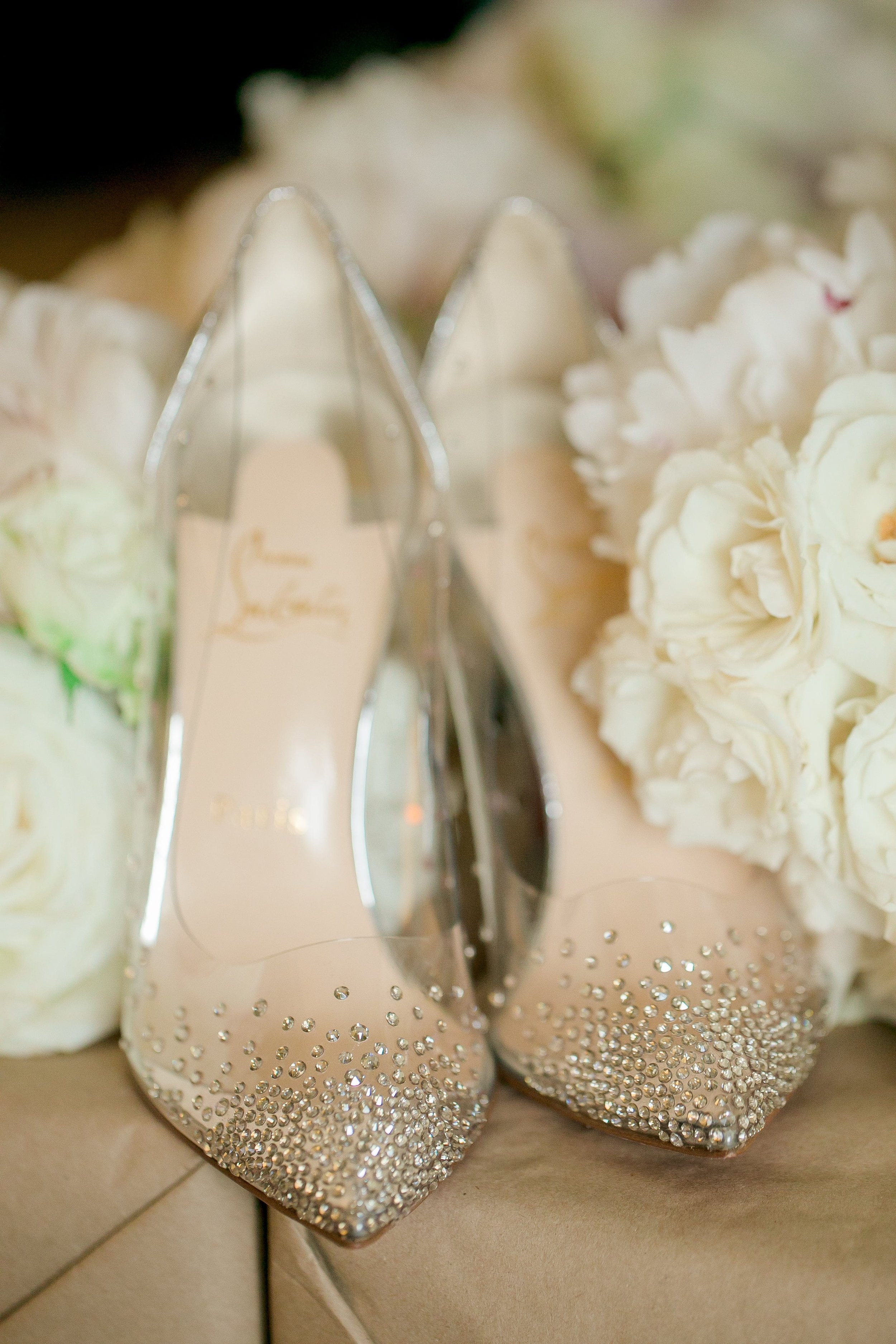 A pair of silver high-heeled shoes decorated with rhinestones on the toes, surrounded by white flowers.