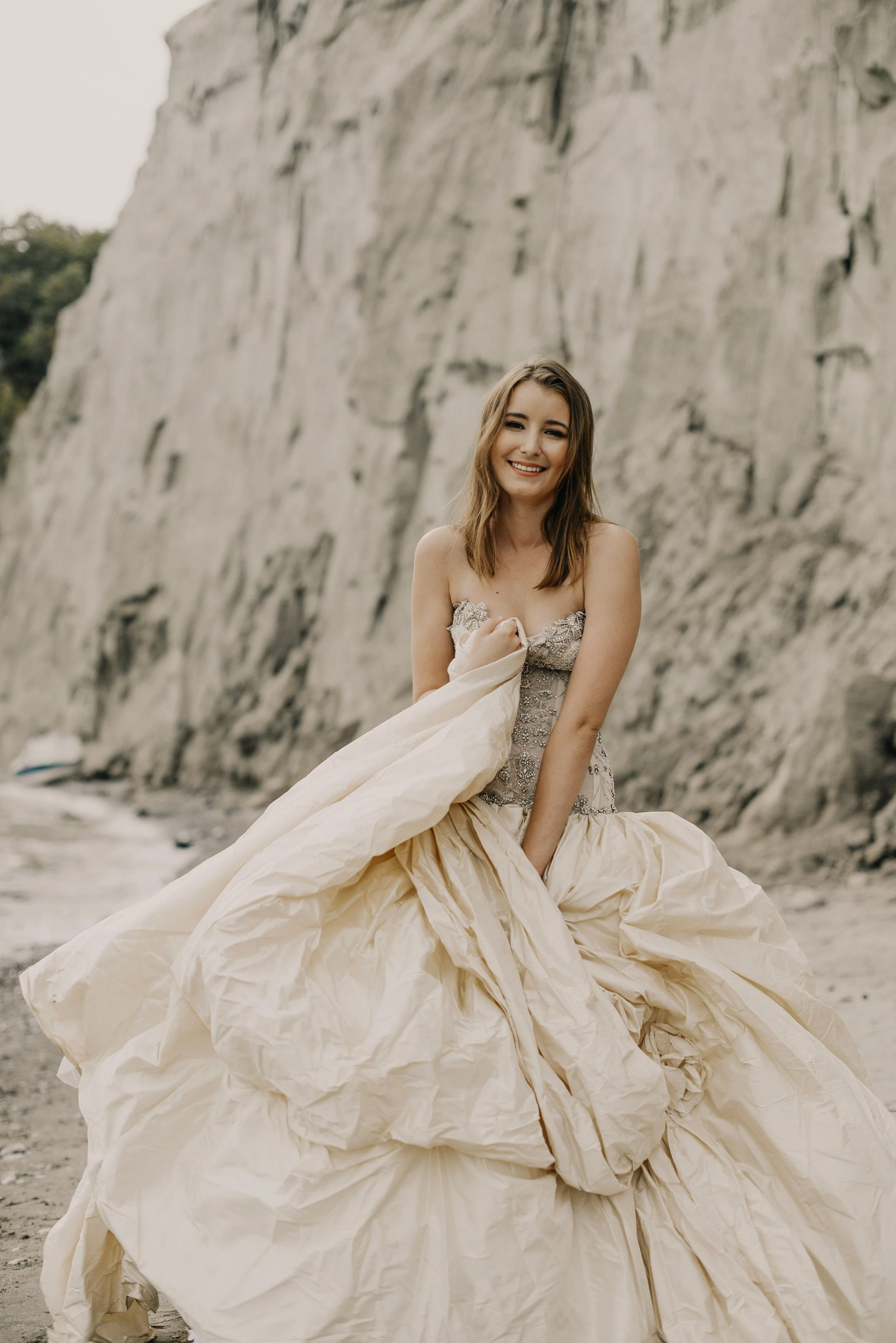A woman in a wedding gown standing on a beach with cliffs in the background, smiling at the camera.
