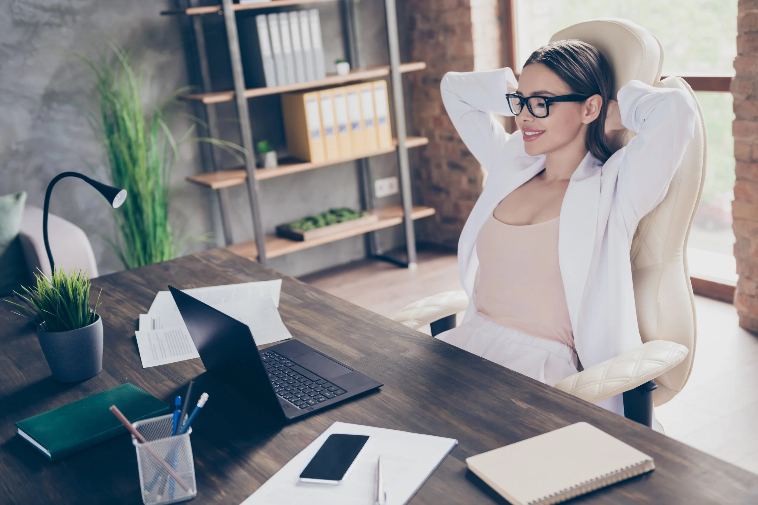 Woman with glasses sitting at a cluttered wooden desk in a modern office, stretching back in a cream-colored chair, with laptop, tablets, notebooks, and office supplies on the desk, and green plants and shelves in the background.
