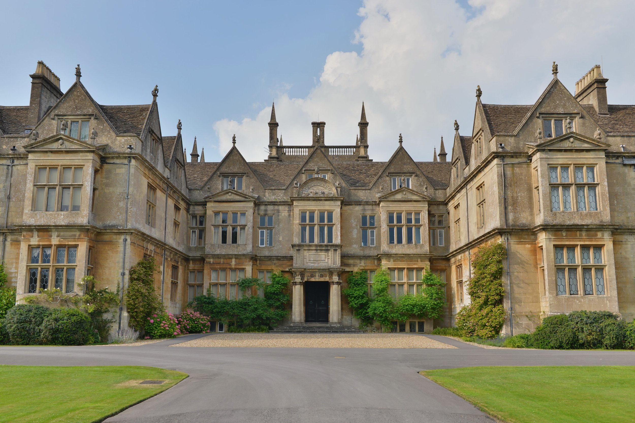 A large, historic castle with multiple turrets, ornate windows, and a grand entrance, surrounded by well-maintained lawns and greenery.