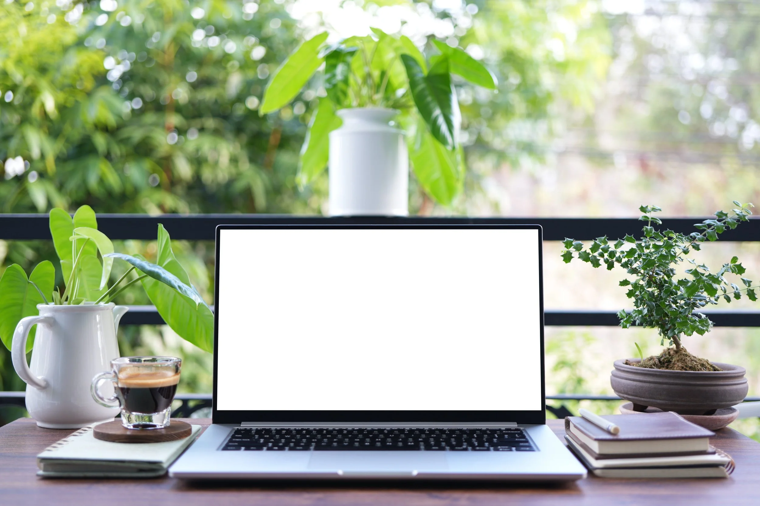 A workspace with a laptop, notebook, and a pen on a wooden table surrounded by potted plants, with a window overlooking greenery outside.