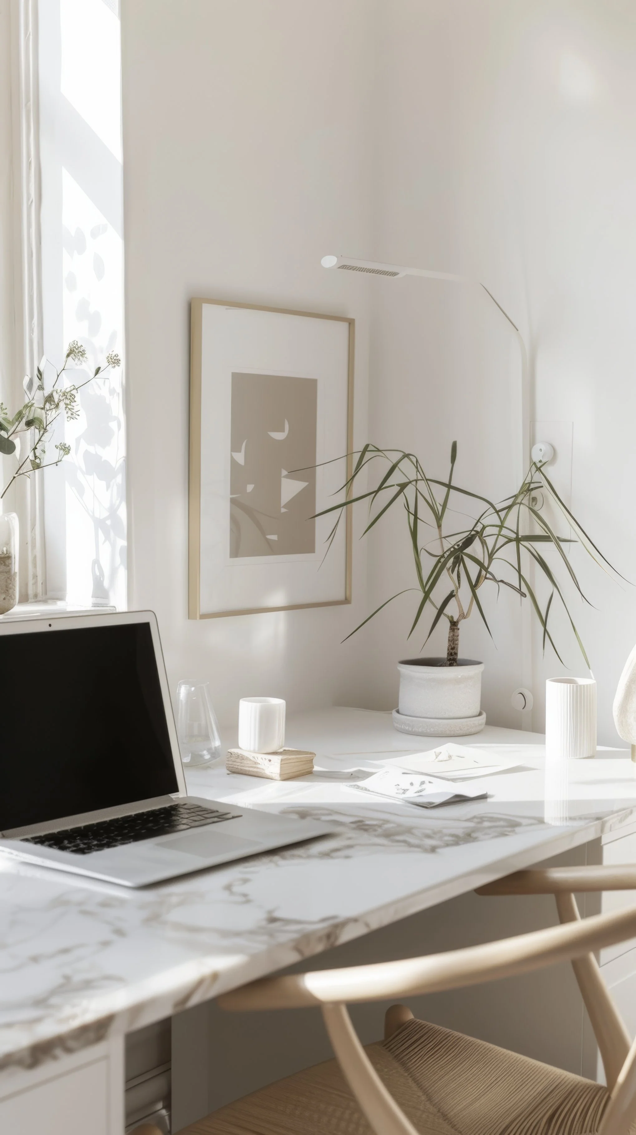 A bright, minimalistic home office with a white marble desk, a laptop, and a potted plant. Decor includes a framed abstract art piece on the wall and table lamps, with natural light coming through a window.
