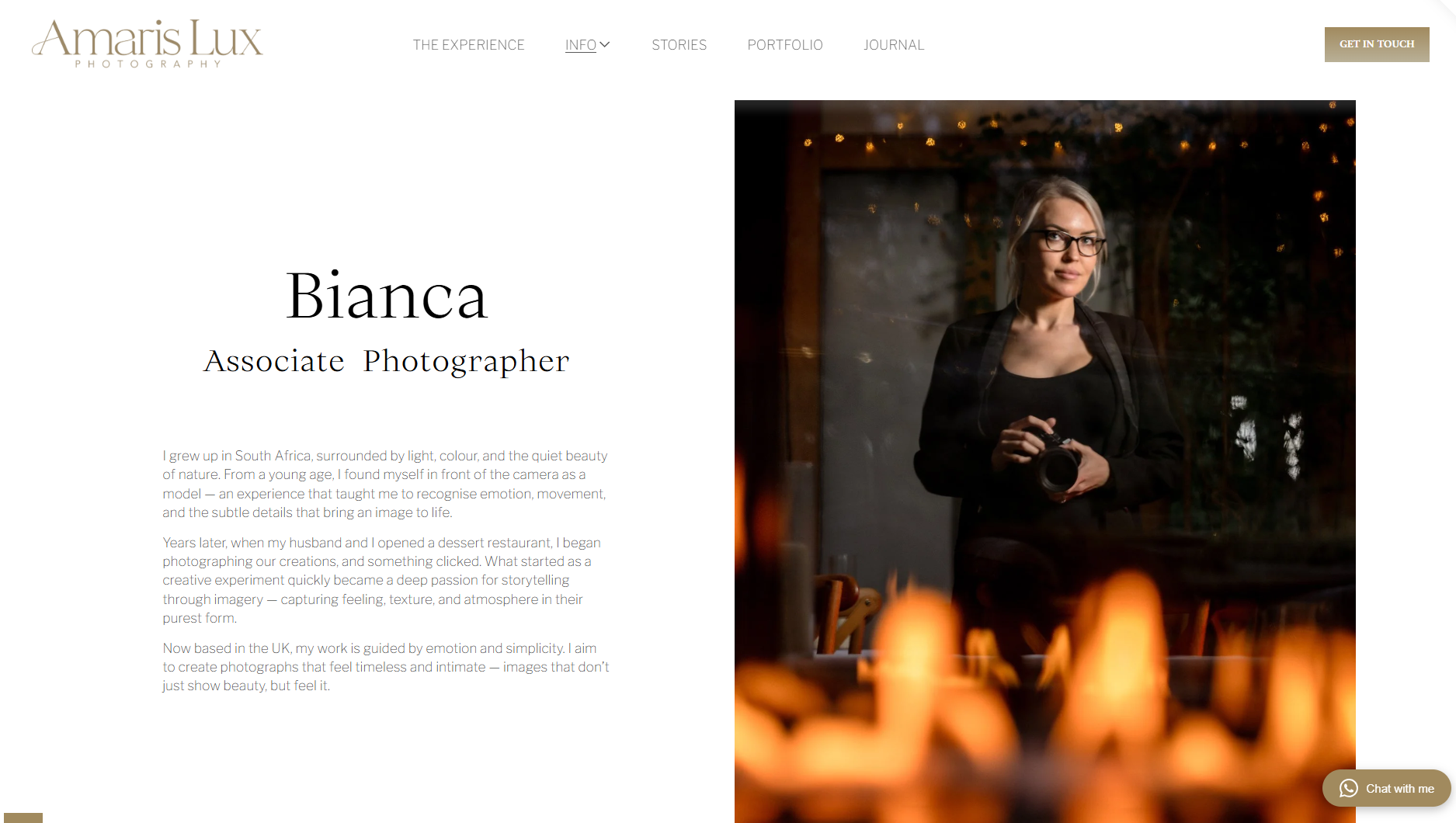 Photographer Bianca standing indoors, holding a camera, with a reflection of a interior setting with warm lights and candles