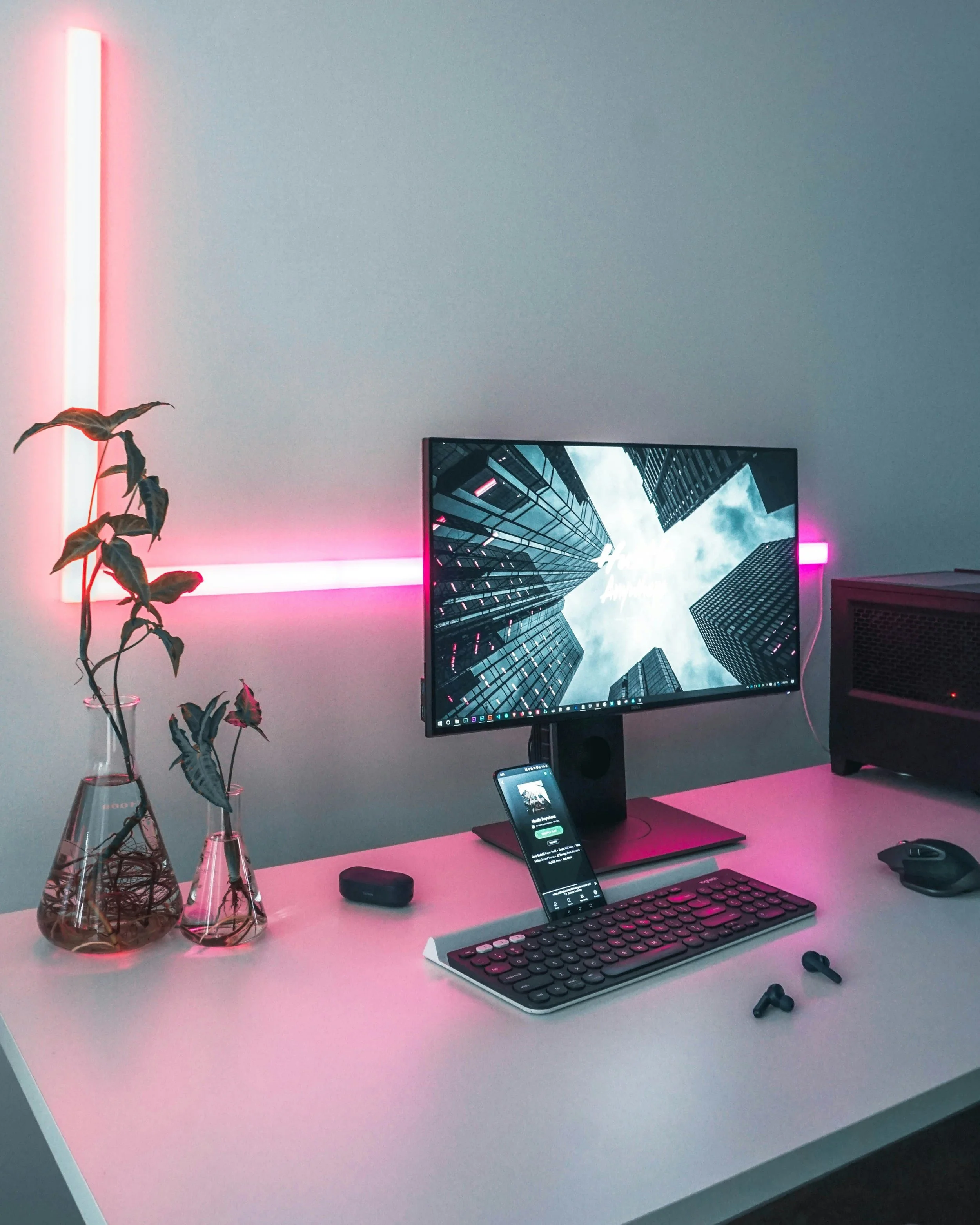 A modern computer workspace featuring a monitor displaying a cityscape, a smartphone with music app, a wireless keyboard, a mouse, wireless earbuds, two vases with leaves, pink neon lights on the wall, and a black computer tower.