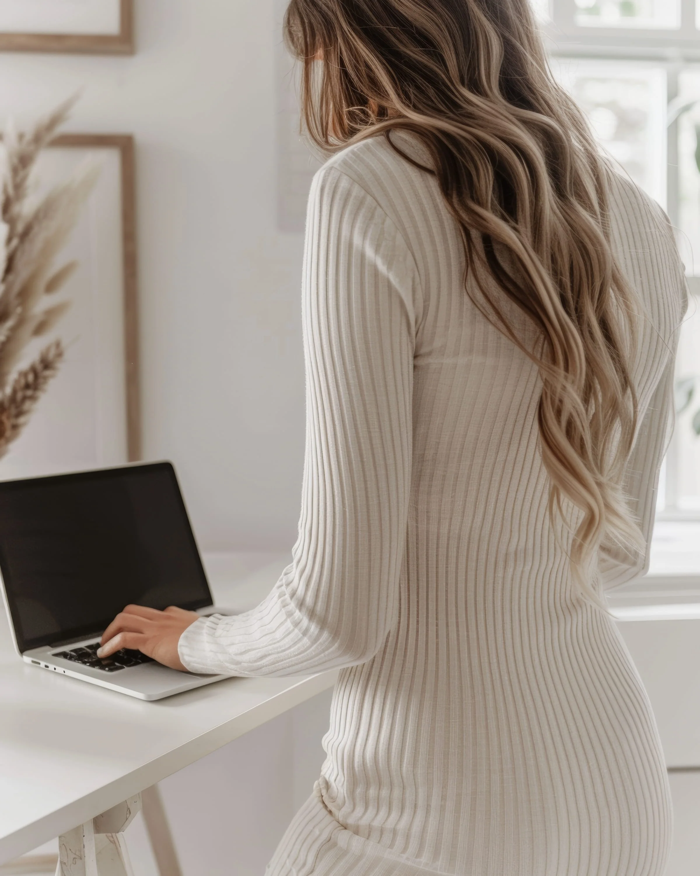 Woman with long wavy hair using a laptop on a white desk in a bright room.