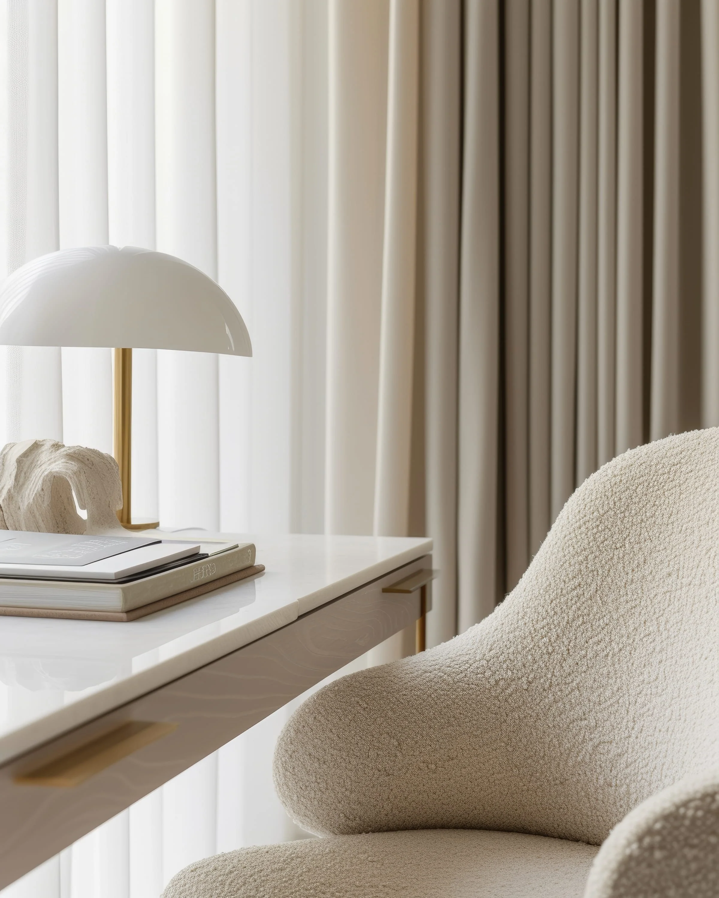 A beige textured armchair beside a white desk with a white and gold desk lamp, a closed book, and a shell-shaped decorative object, with beige curtains in the background.