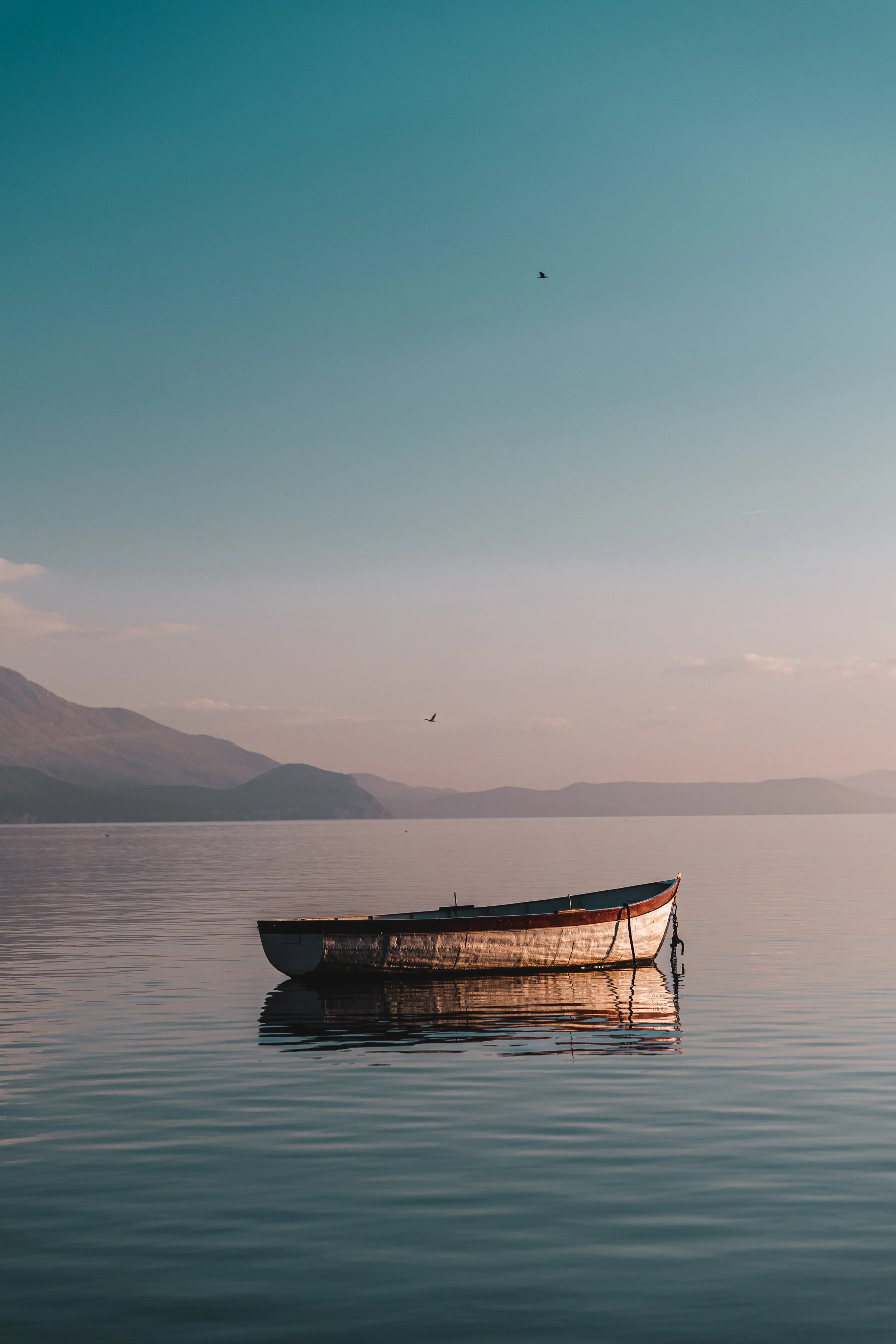 A solitary boat floating on a calm lake with mountains in the background and birds flying in the sky.