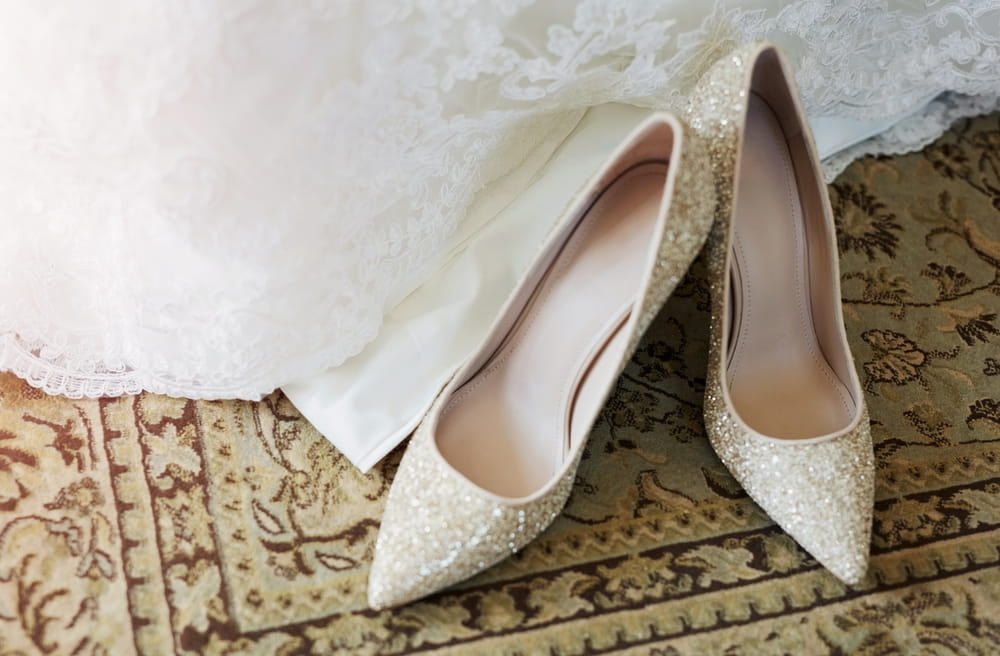 A pair of white, glittery high-heeled shoes resting on an ornate, patterned carpet, partially obscured by a white and lace dress.