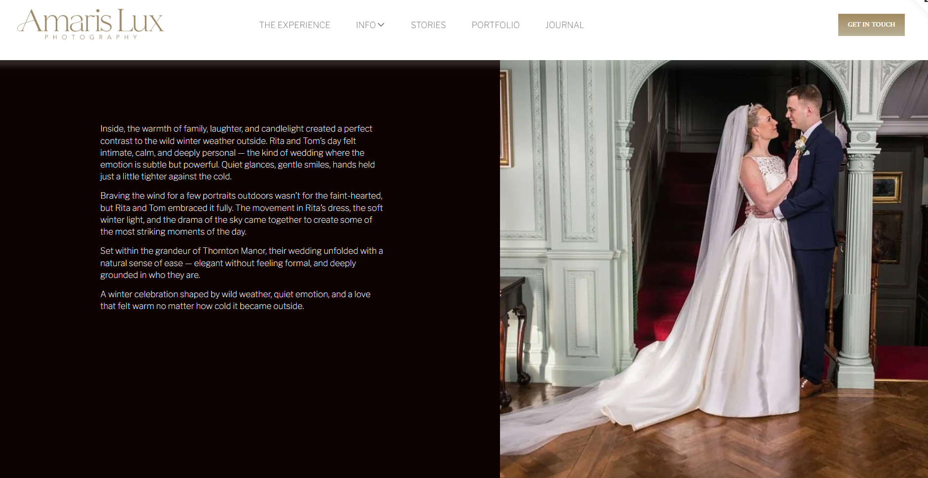 A bride and groom stand close together inside a grand, ornate interior. The bride is wearing a white wedding gown with lace details and a long veil, and the groom is dressed in a navy blue suit with a boutonnière. They are gazing into each other's eyes, touching hands, with a staircase and framed artwork in the background.