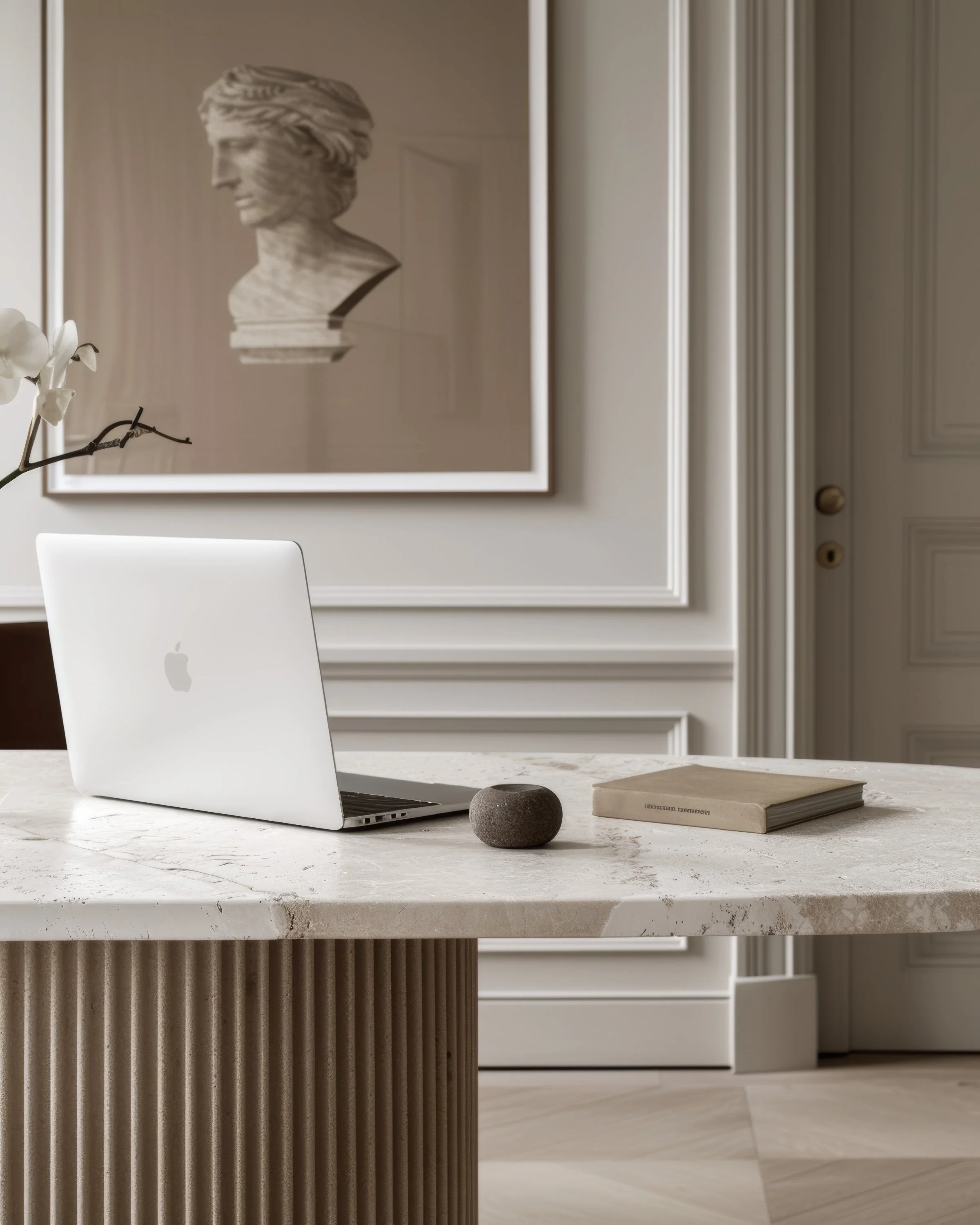 Modern interior with a marble kitchen island, a white orchid, a closed laptop with a logo, a small stone, and a closed book on the countertop. A framed artwork of a classical bust hangs on the wall.