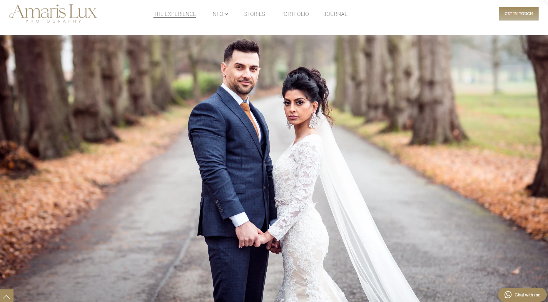 A bride and groom holding hands, standing on a tree-lined path outdoors, dressed in wedding attire with the bride in a lace wedding gown and the groom in a suit.