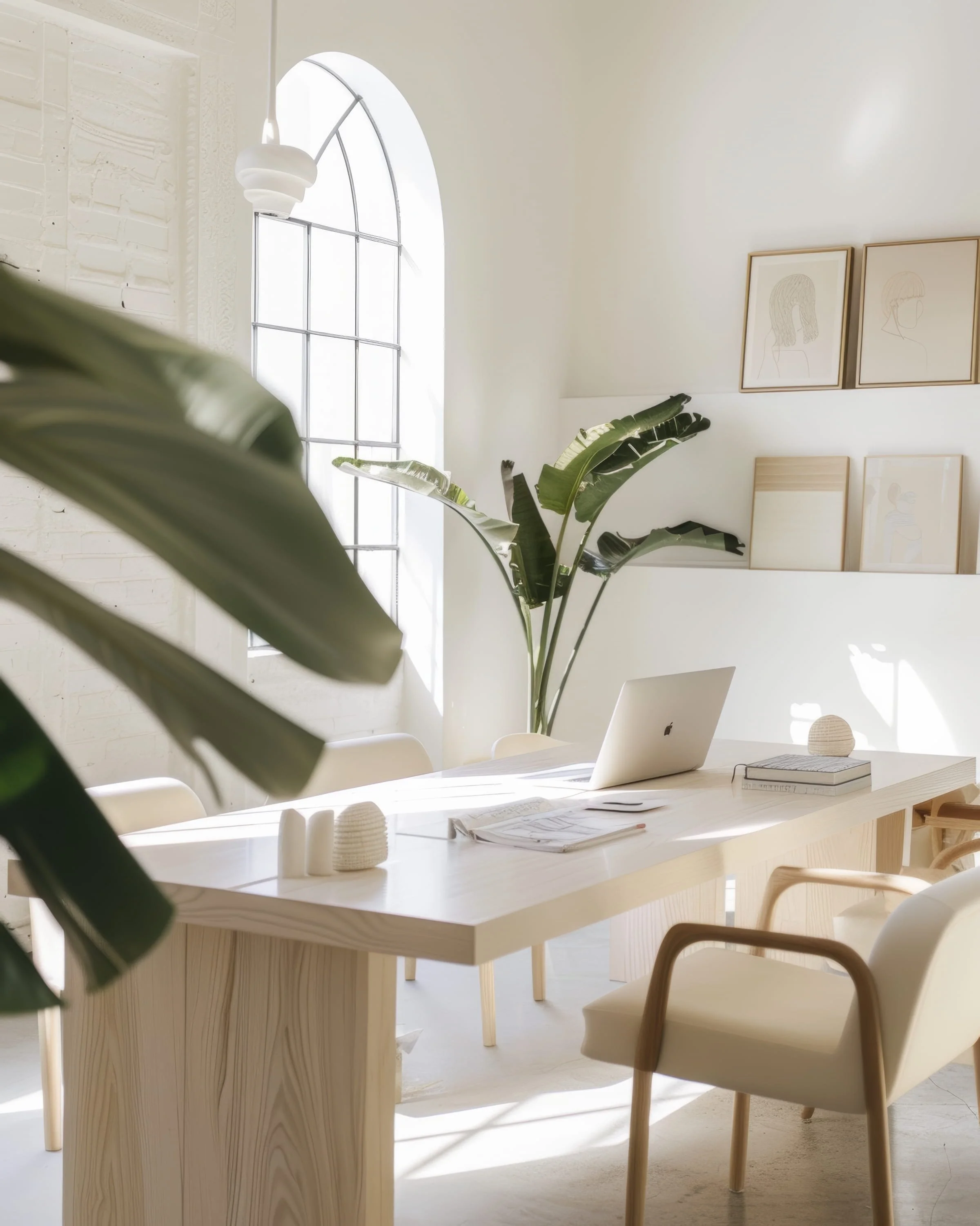 A bright, minimalist office space with large arched window allowing natural light. A white desk with a laptop, notebooks, and decorative objects. Light-colored chairs, framed line art on the wall, and green potted plant add aesthetic touch.