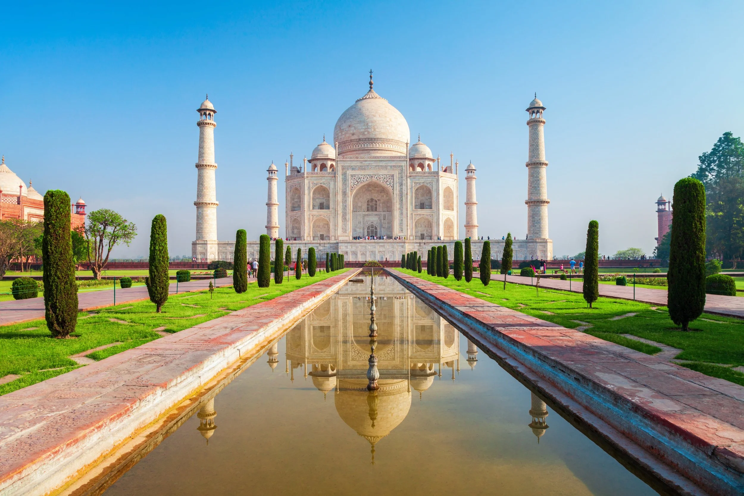 The Taj Mahal, a white marble mausoleum with four minarets, surrounded by lush green gardens and a reflecting pool, under clear blue sky.