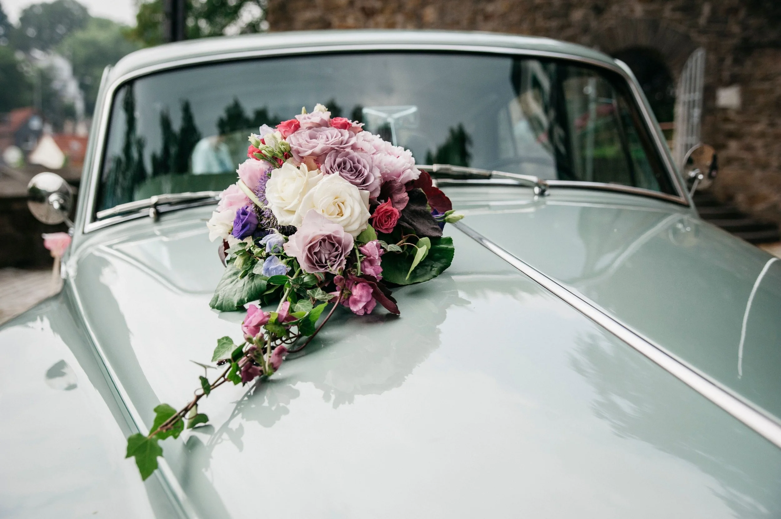 A vintage green car with a floral bouquet of pink, purple, and white flowers on the hood.