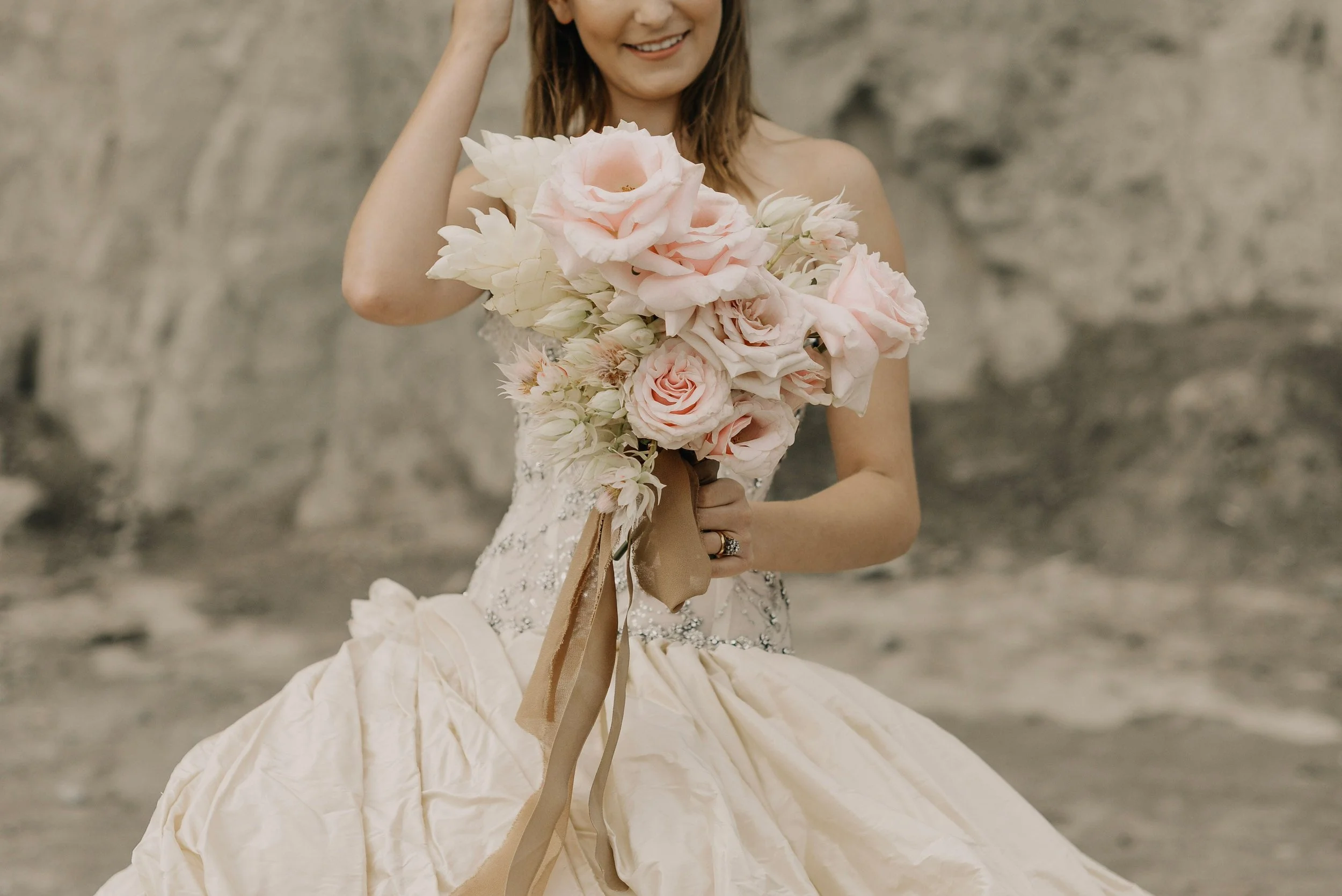 A woman in a wedding gown holding a large bouquet of pink and white roses and lilies outdoors near a rocky background.