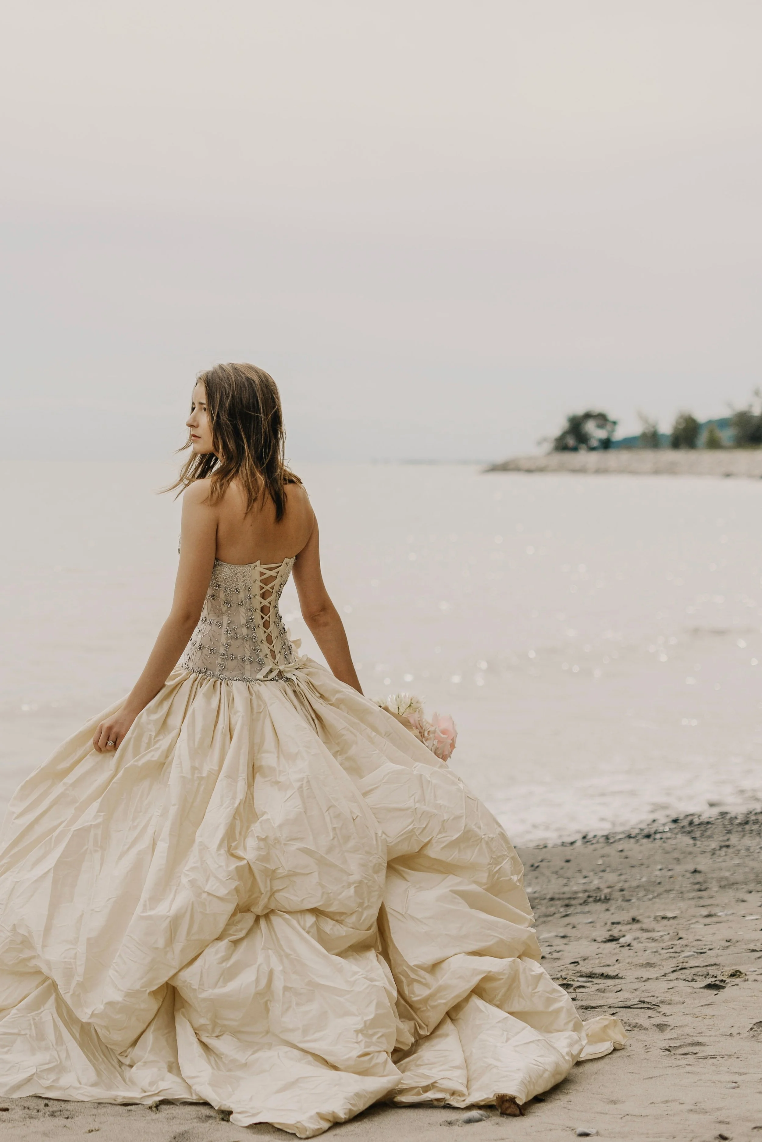 A woman in an elaborate wedding dress with a corset back, holding a bouquet, standing on a beach near the water under a cloudy sky.