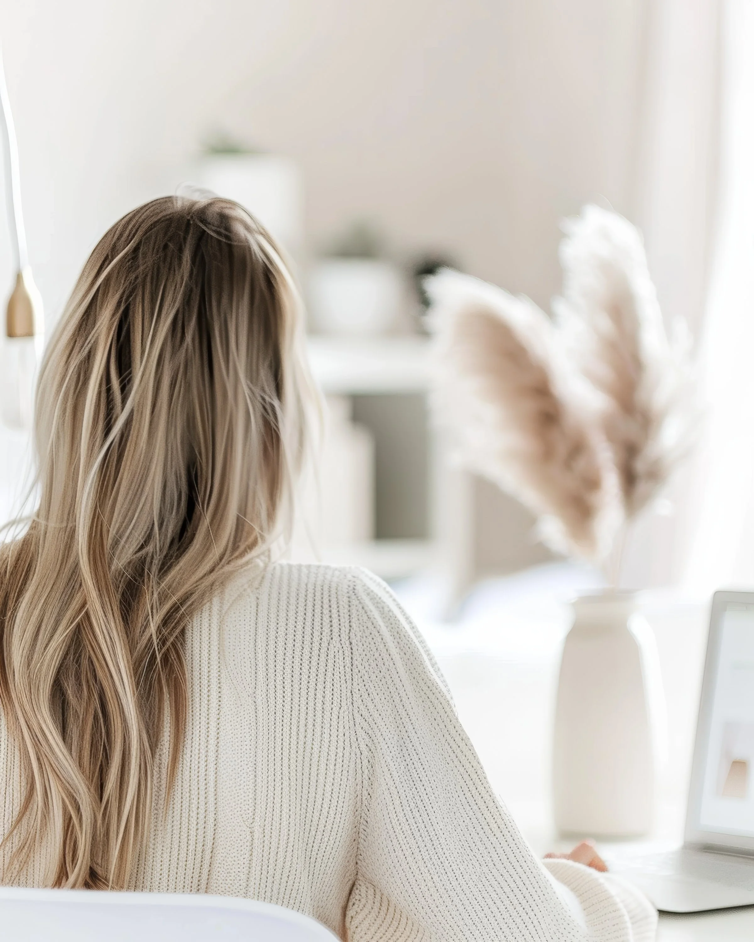 Back view of a woman with long blonde hair working on a laptop at a white desk in a bright, modern room with neutral decor.