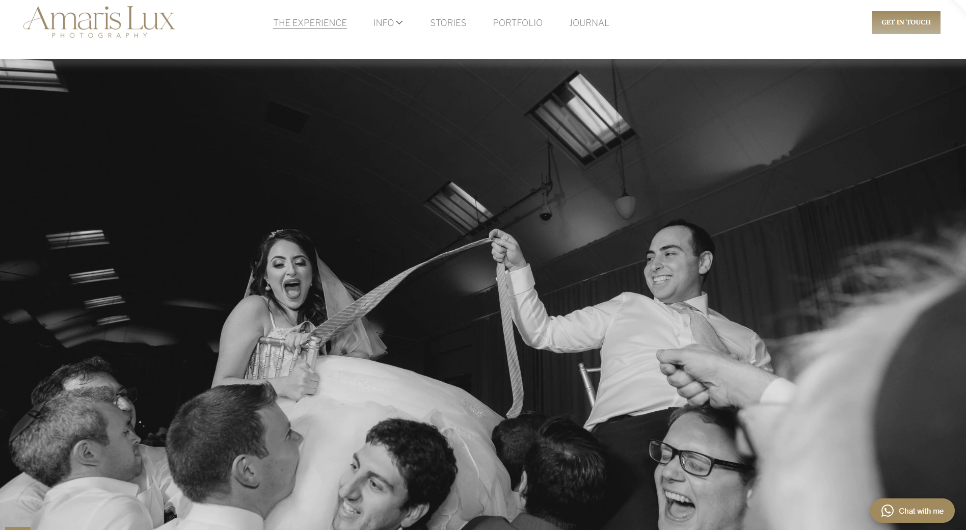 Black and white photo of a wedding reception with the bride and groom being lifted on a chair by guests; the bride is holding a ribbon and appears excited, while the groom is smiling. The scene is lively with people around.