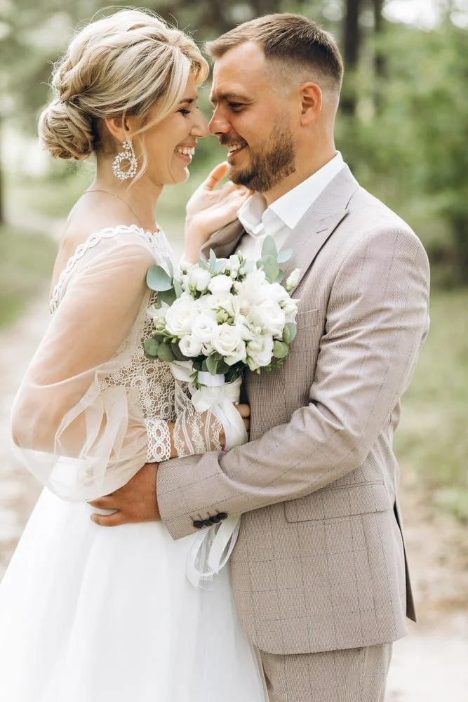 A bride and groom smiling and looking into each other's eyes outdoors, holding a bouquet of white flowers.