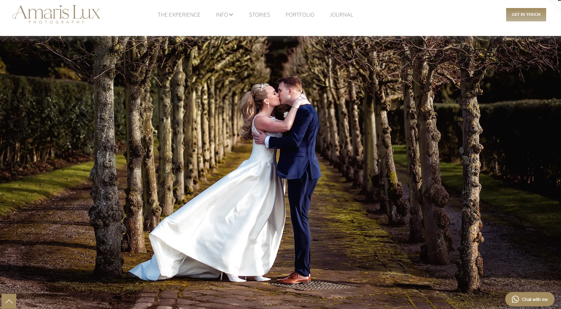 A bride and groom kissing in a tree-lined outdoor pathway during their wedding, with the bride in a white gown and the groom in a dark suit.